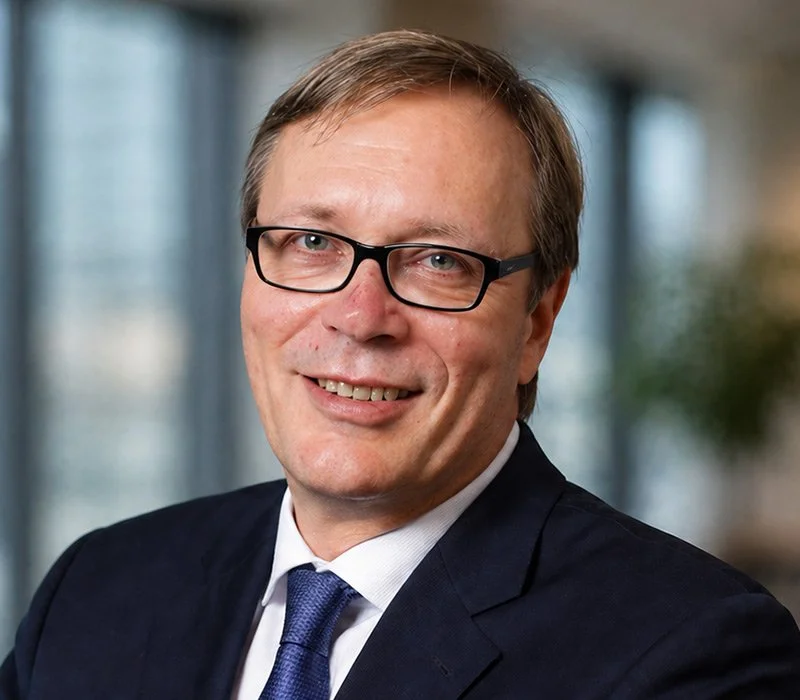 Porträt eines Mannes mit Brille in formellem Anzug und Krawatte, lächelnd, vor Büro-Hintergrund.