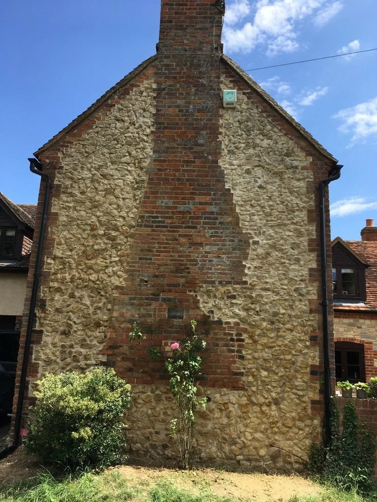 A brick church wall with a vertical strip of bricks in the center, partially covered by beige and yellowish stonework. There is a small plant with pink flowers in front of the wall, and a bush to the left. The sky is blue with some clouds.
