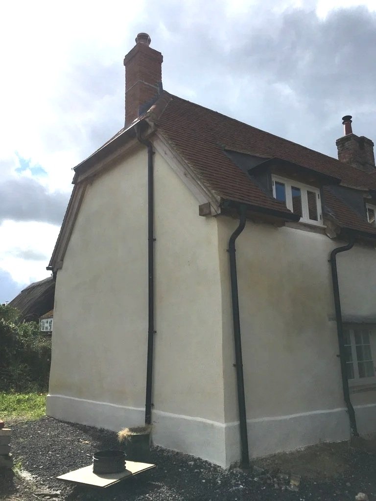Side view of a building showing finished Lime restoration with a sloped roof, chimney, and black rain gutters, with a cloudy sky in the background.