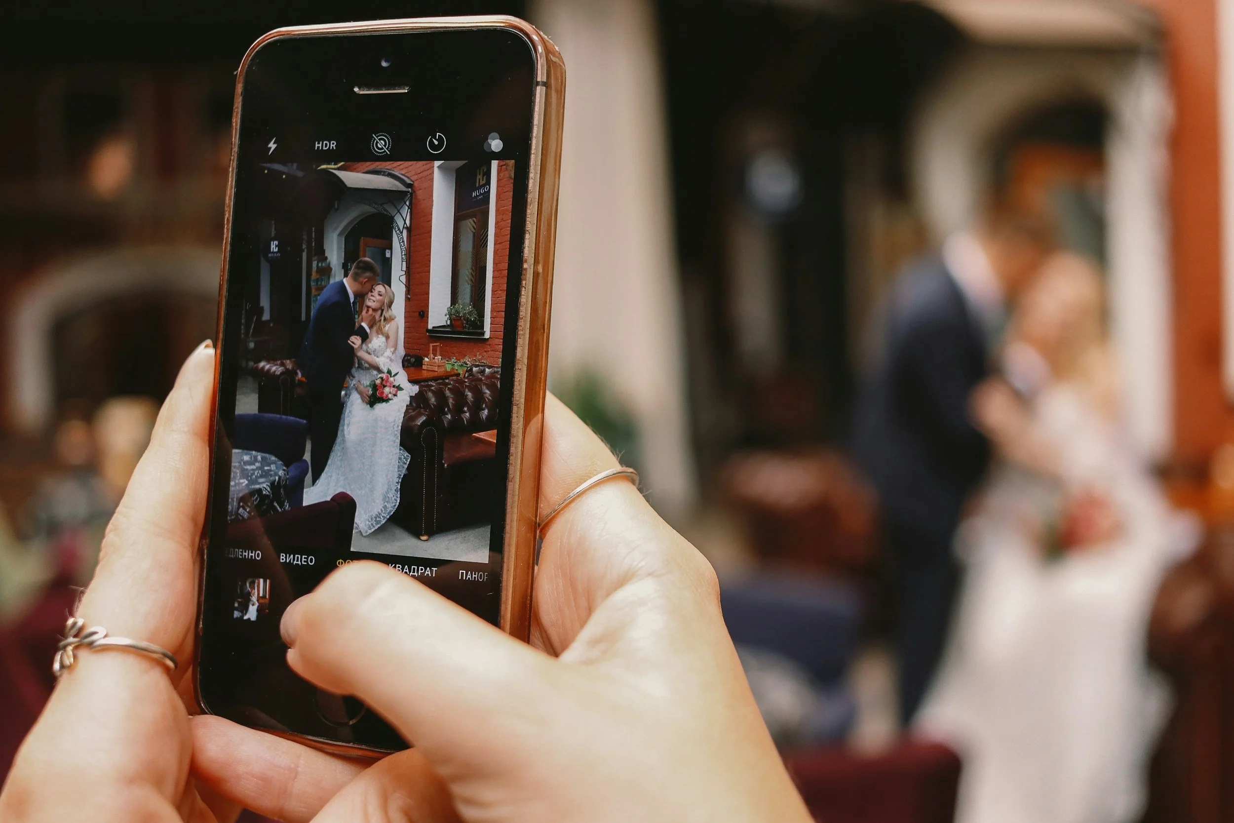 Close-up of a person holding a smartphone taking a photo of a couple in wedding attire inside a cozy, stylish venue with brick and white walls and large windows.