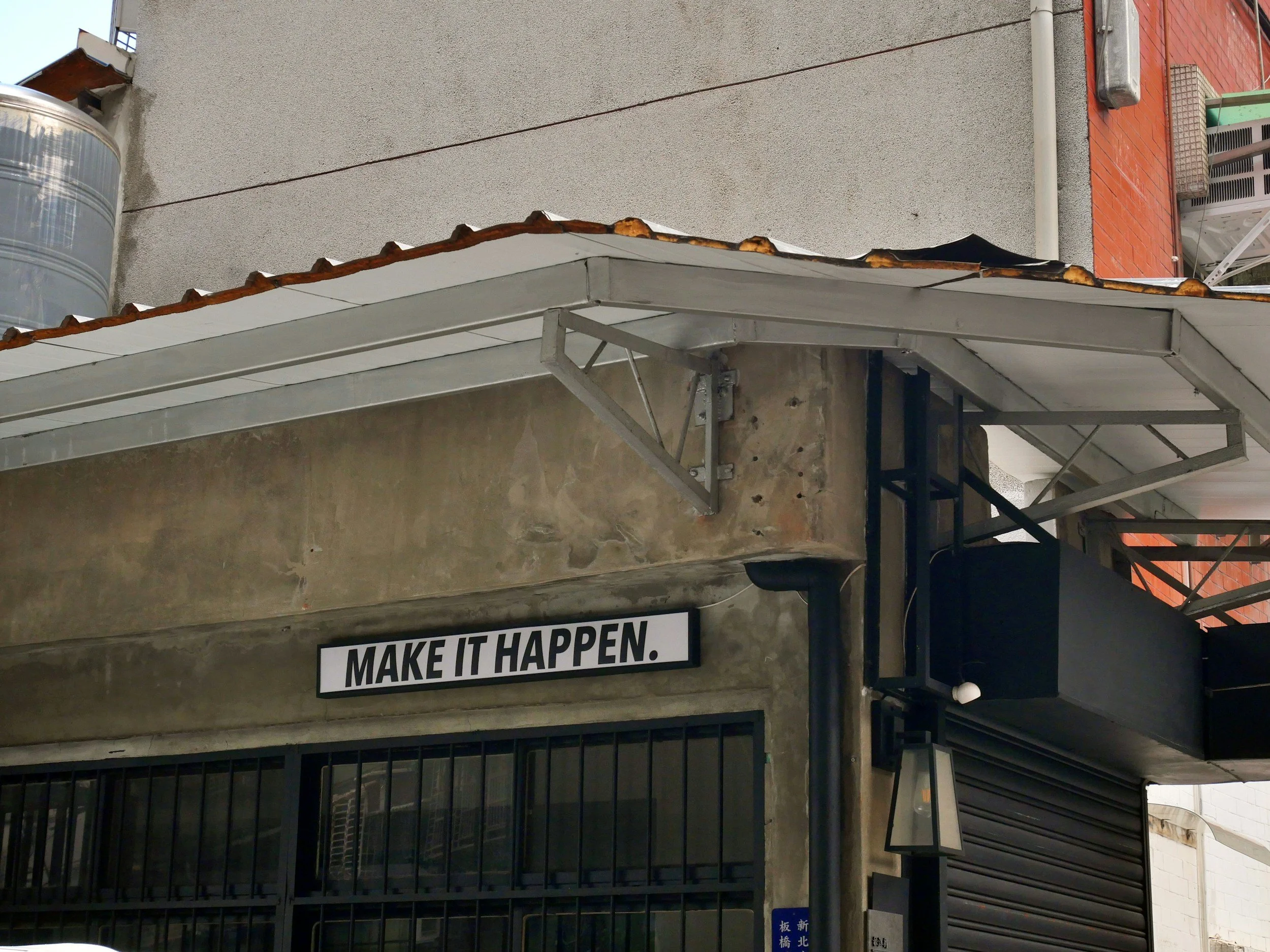 A building with a sign that reads "MAKE IT HAPPEN." in black and white, underneath a metal canopy with a tiled roof. There are stairs and a gated entrance beneath the sign.