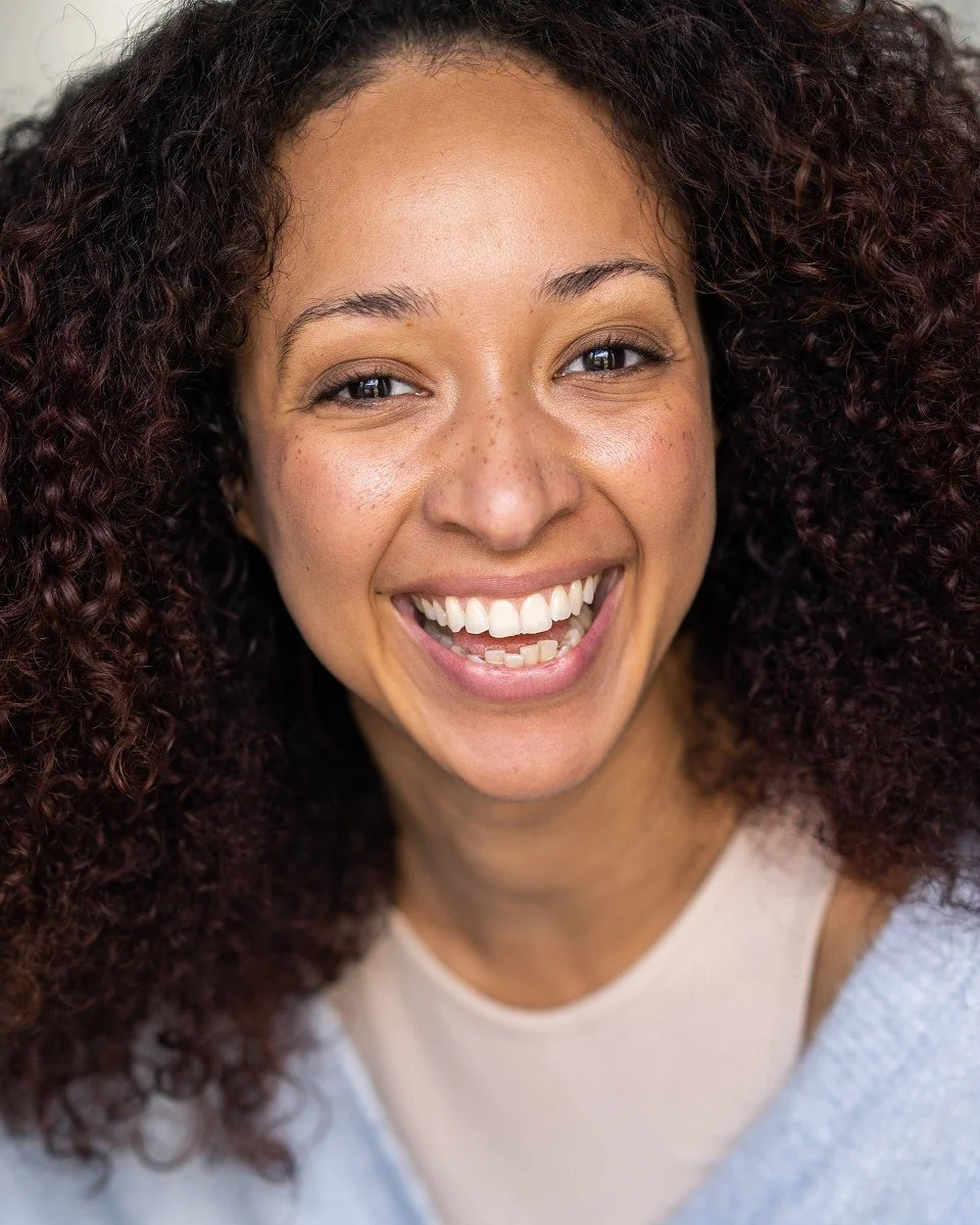 Close-up of a woman with curly dark hair smiling brightly, showing her teeth, wearing a white top.