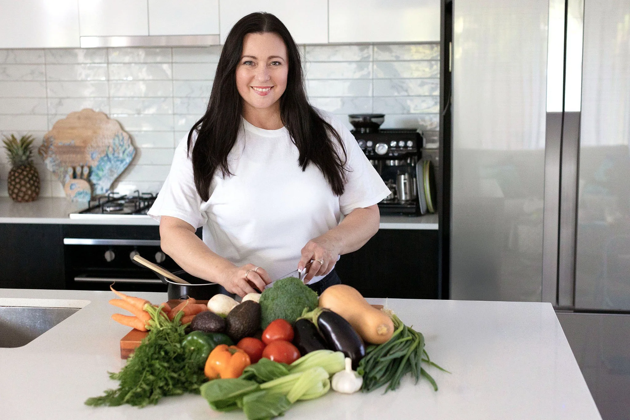 Woman with long dark hair preparing vegetables in a modern kitchen with a white tile backsplash and stainless steel appliances, vegetables on counter including carrots, tomatoes, broccoli, eggplant, green onions, garlic, and avocado.