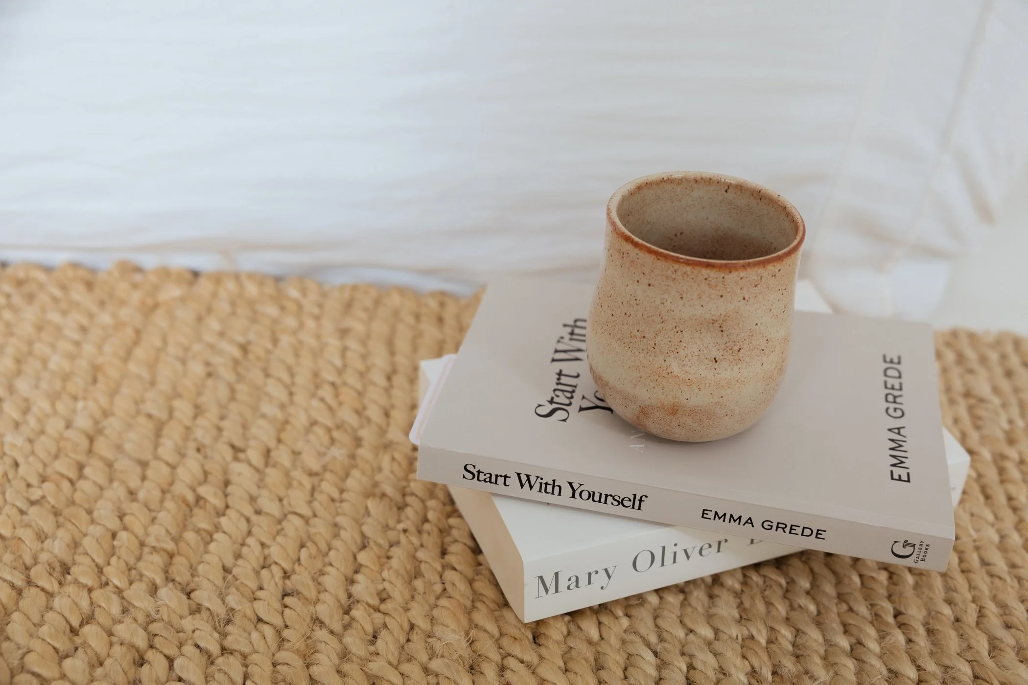 A stack of three books with titles including "Start With Yourself" and author names Emma Gredé and Mary Oliver, with a speckled beige ceramic cup placed on top of the books on a textured woven surface.