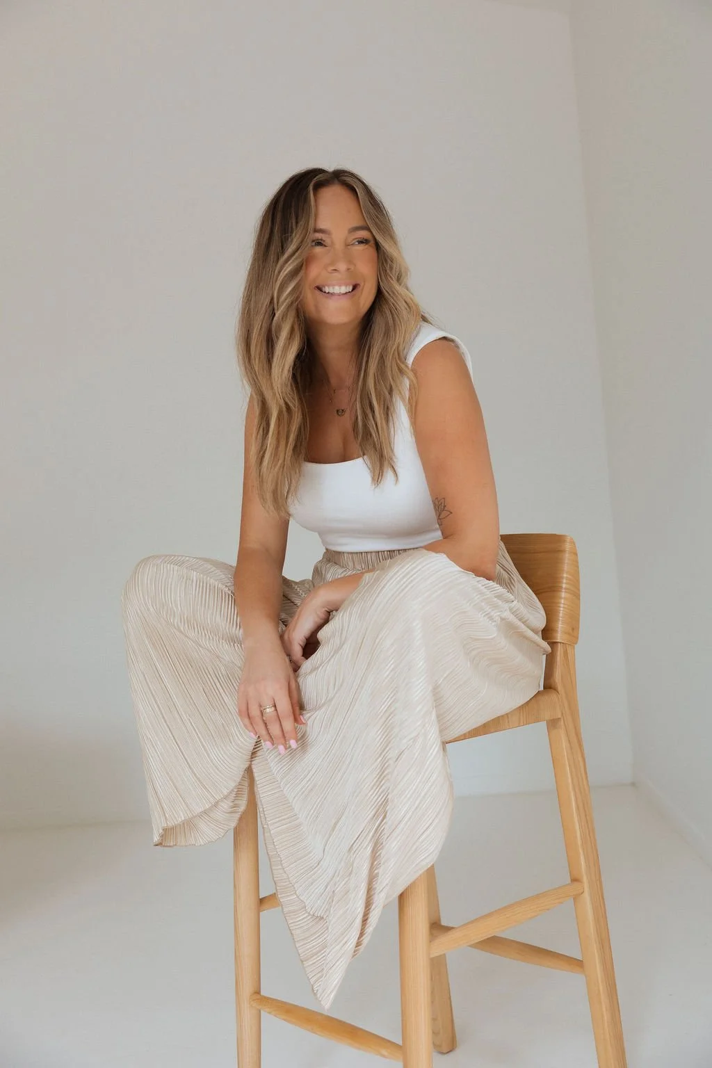 A woman with wavy, shoulder-length hair, wearing a white sleeveless top and a beige pleated skirt, sitting on a wooden chair and smiling in a minimalistic, light-colored room.