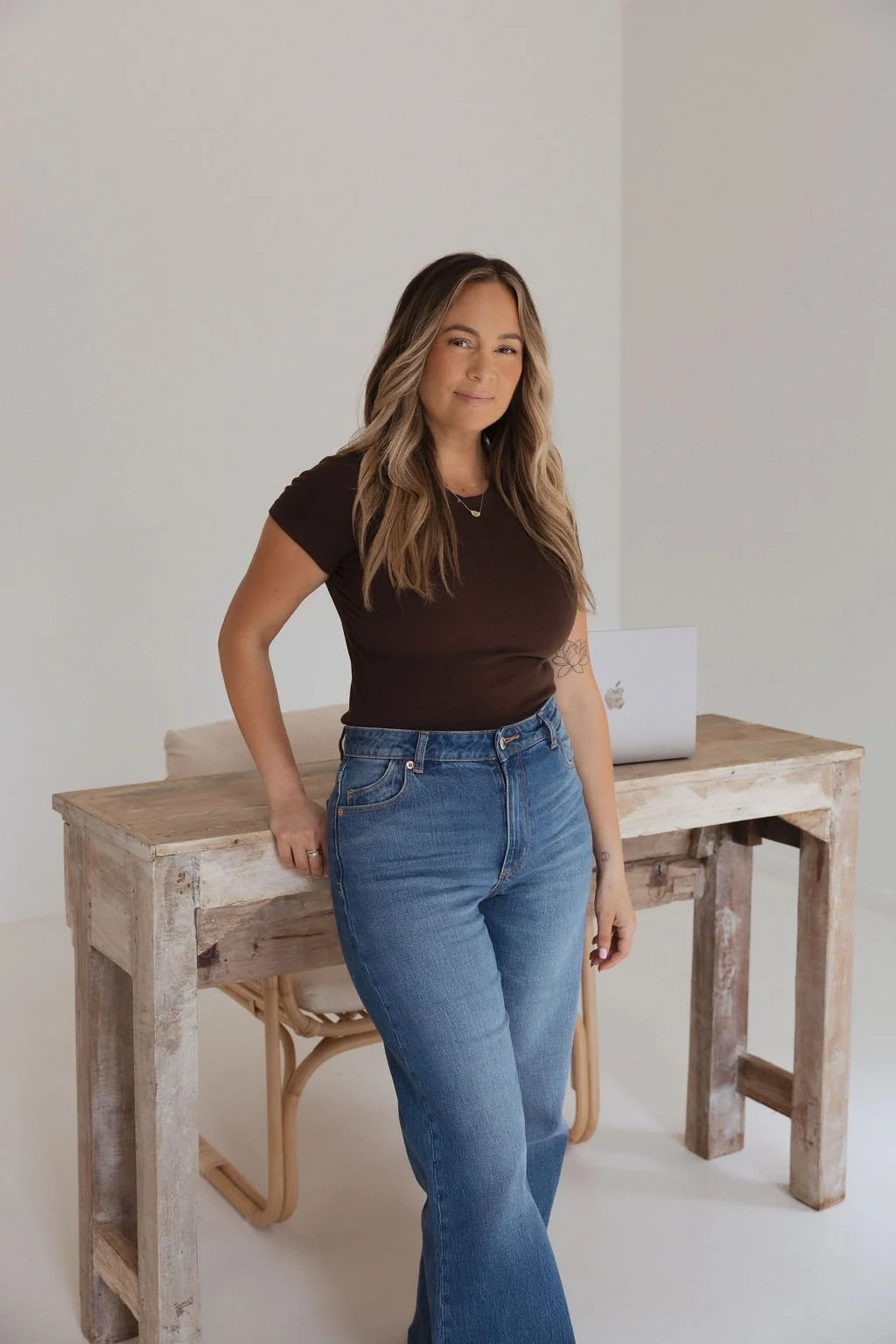 A woman with long wavy hair, wearing a black t-shirt and blue jeans, standing in front of a wooden desk with a MacBook on it, in a minimalistic room with white walls.