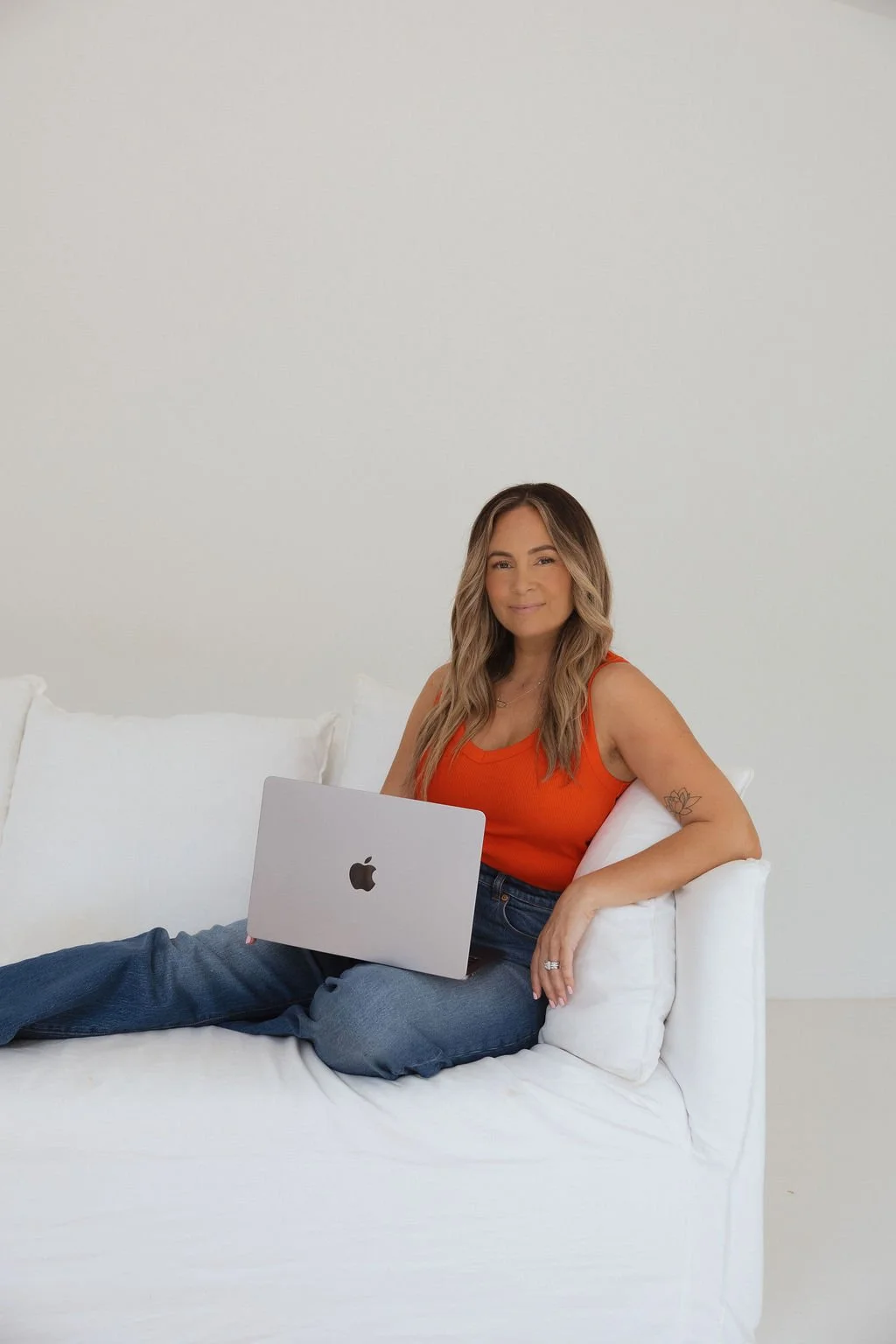Woman sitting on white couch with a laptop on her lap, wearing an orange sleeveless top and blue jeans, against a plain white wall.