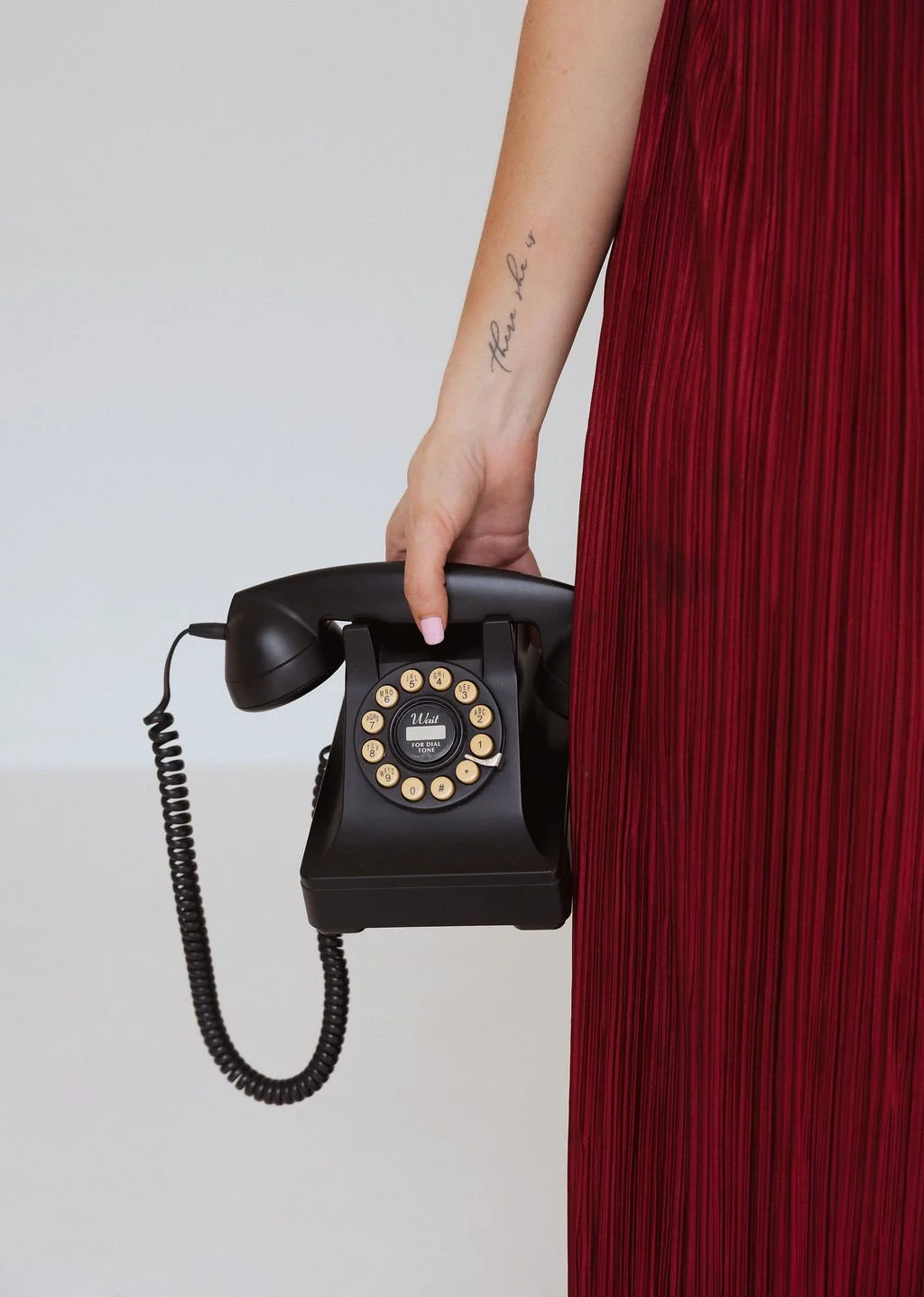 A person in a red dress holds a black vintage rotary phone with a coiled cord, with the phrase "take it" tattooed on their forearm.