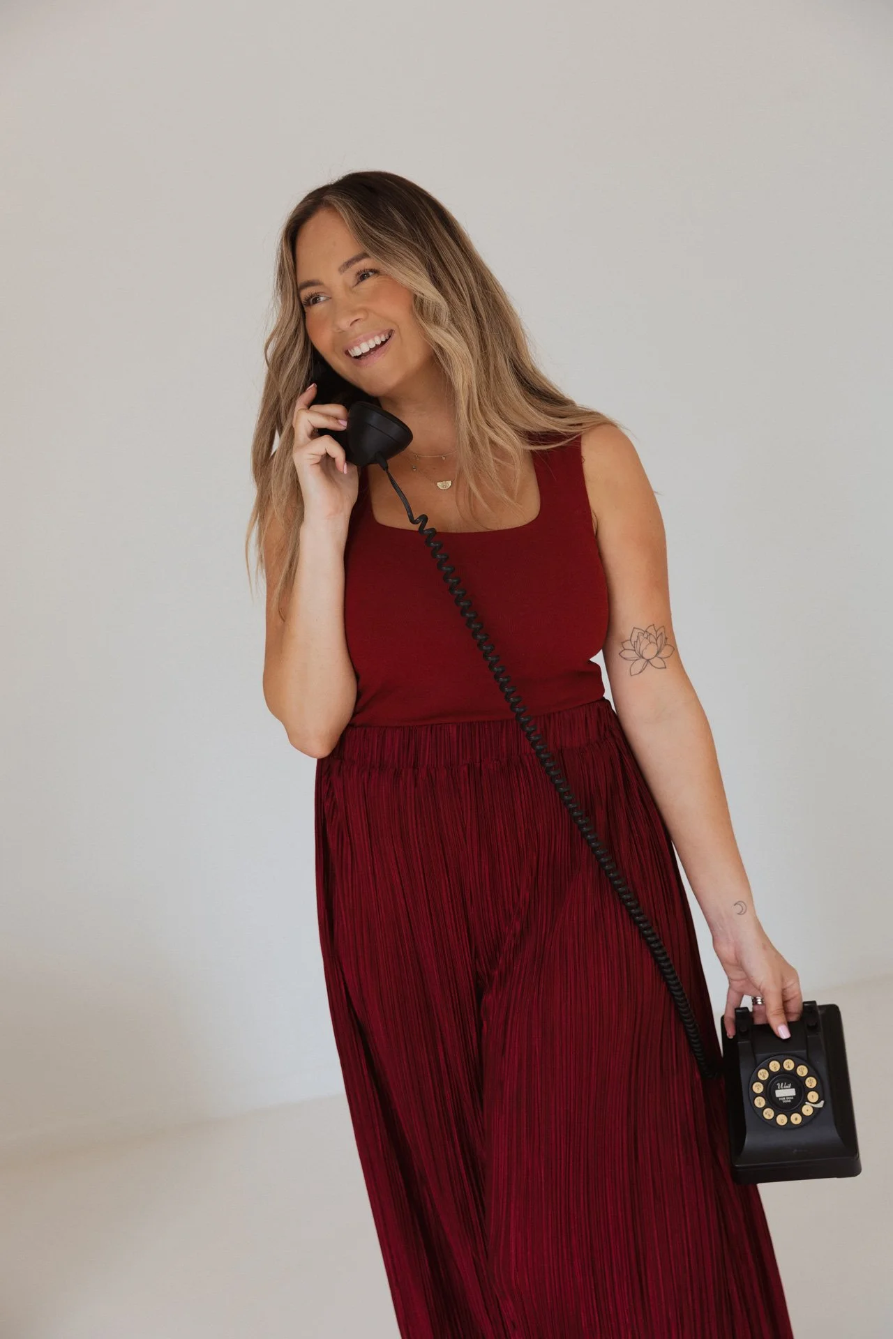 A woman in a red dress winks while talking on a black rotary phone and holding a vintage black rotary dial telephone receiver in her other hand.