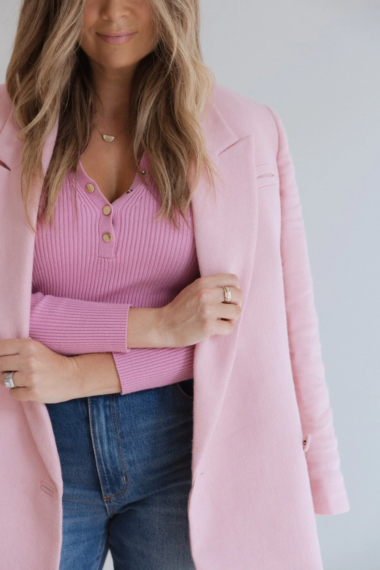 A woman wearing a pink cardigan and top with gold buttons, jeans, and adorned with jewelry including rings and a necklace, standing against a plain background.