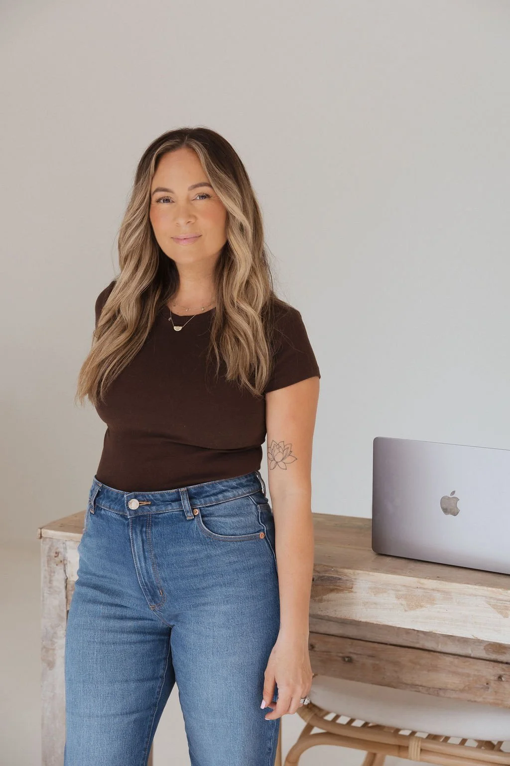 A woman with long, wavy hair and a tattoo of a lotus flower on her left arm, wearing a black t-shirt and blue jeans, standing next to a wooden table with a MacBook laptop.