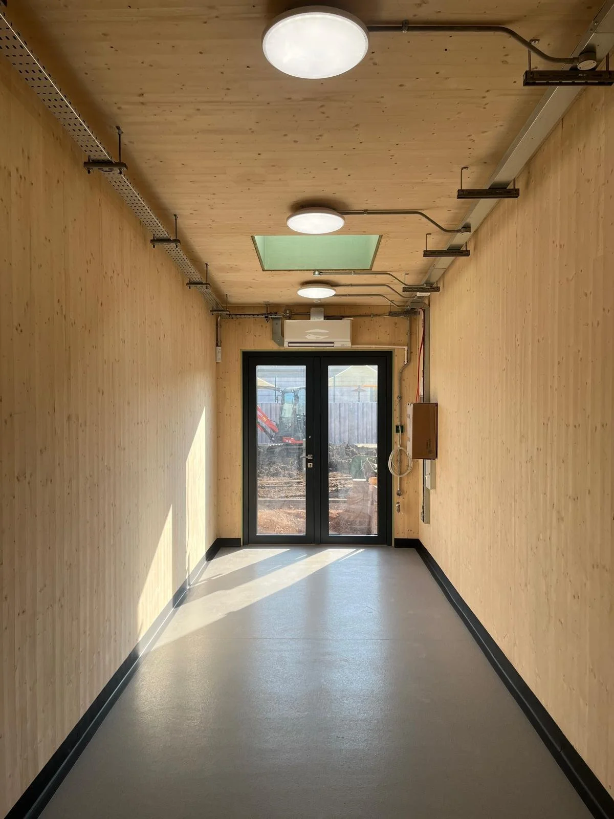 Empty hallway with wood-paneled walls, ceiling, and black-trimmed glass door at the end, showing a construction site outside.