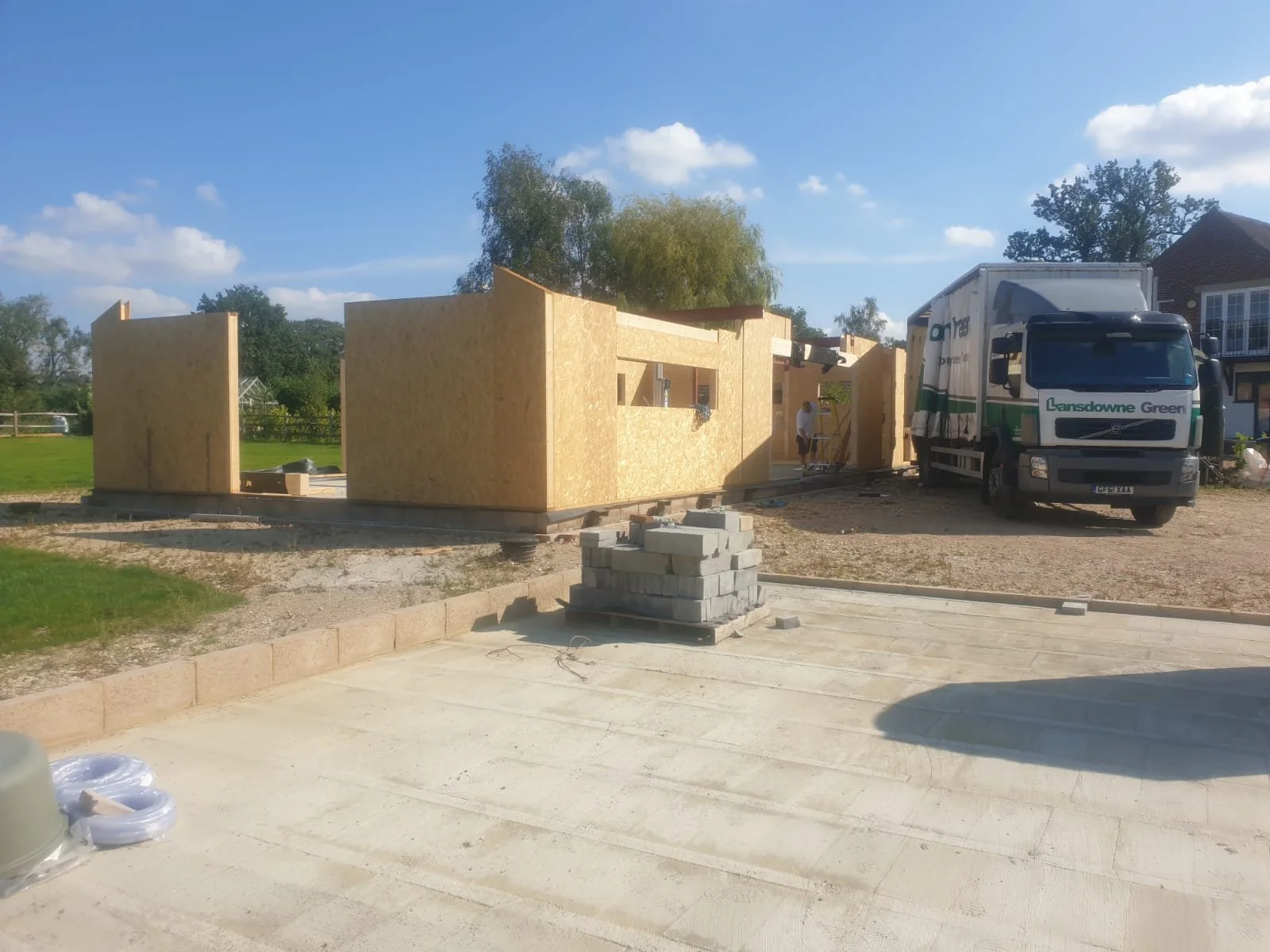 Construction site with wooden walls, a small stack of concrete blocks, and a truck labeled ''Lansdowne Green.'' A person is working near the wooden structure, with a sunny sky and some trees in the background.