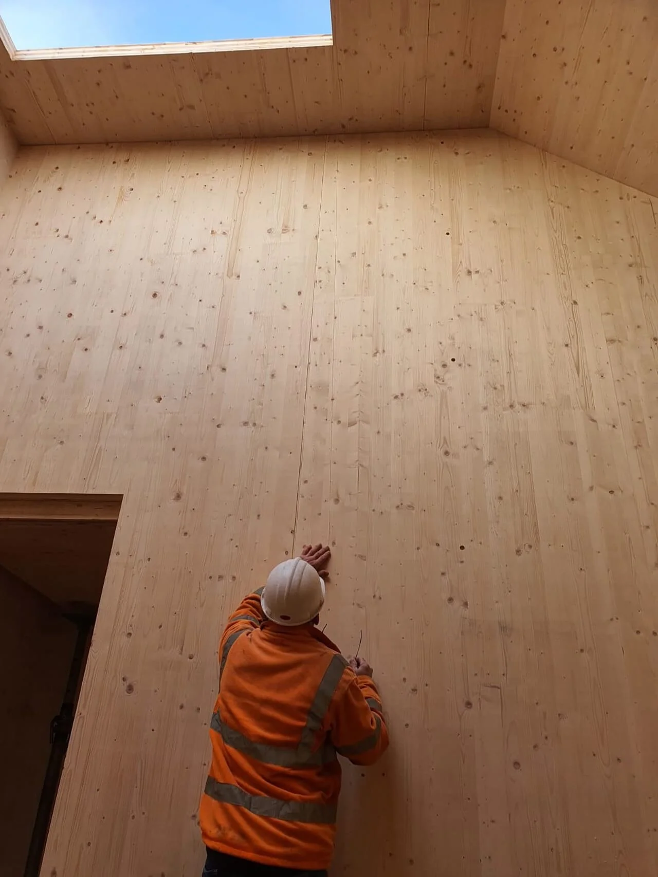 Worker wearing an orange safety jacket and white helmet working on a large wooden wall or ceiling in an indoor construction site.