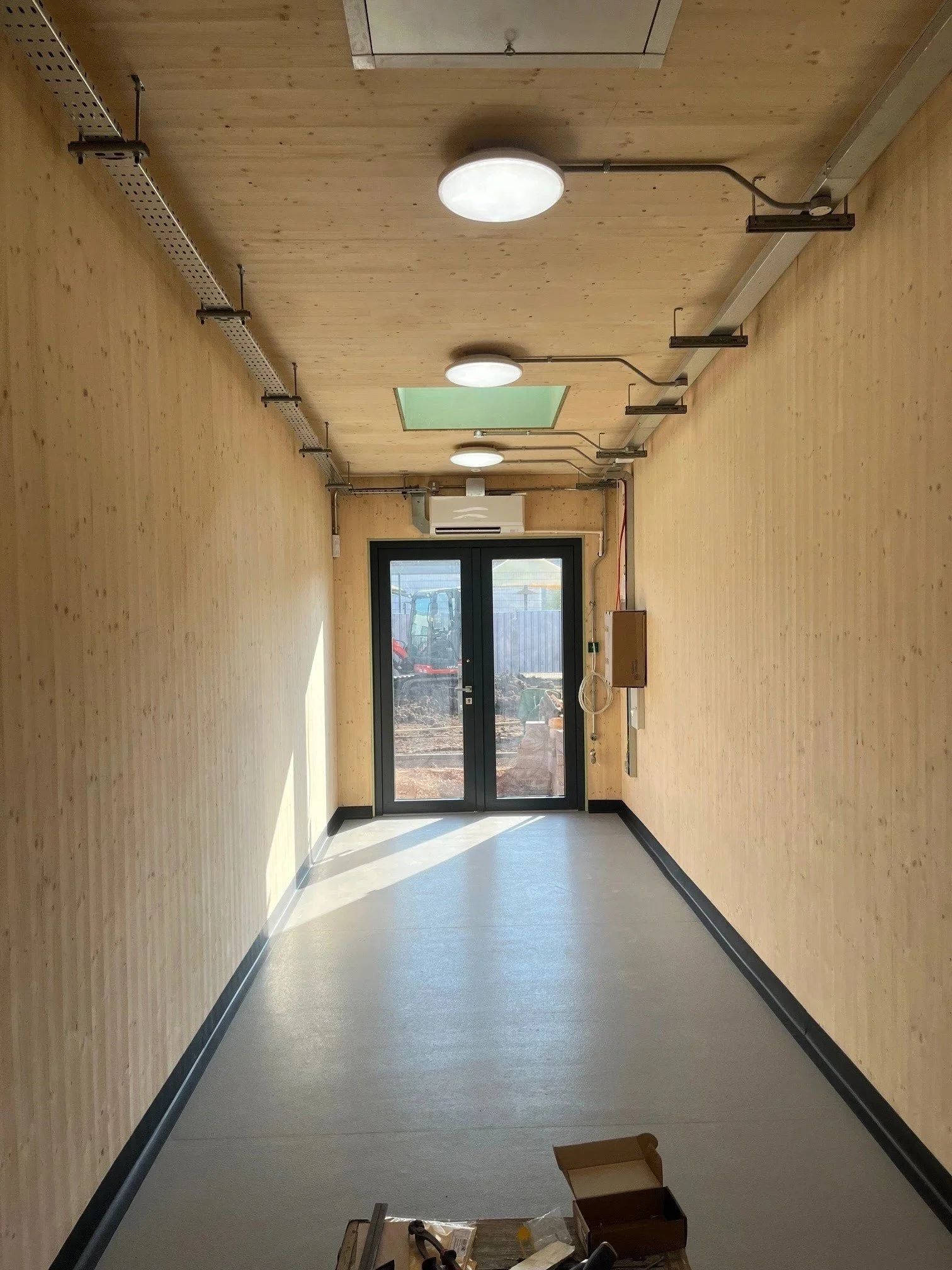 Empty room with wooden walls and flooring, large glass door leading outside, space for installation of equipment or furniture, minimal objects including boxes and a small table.
