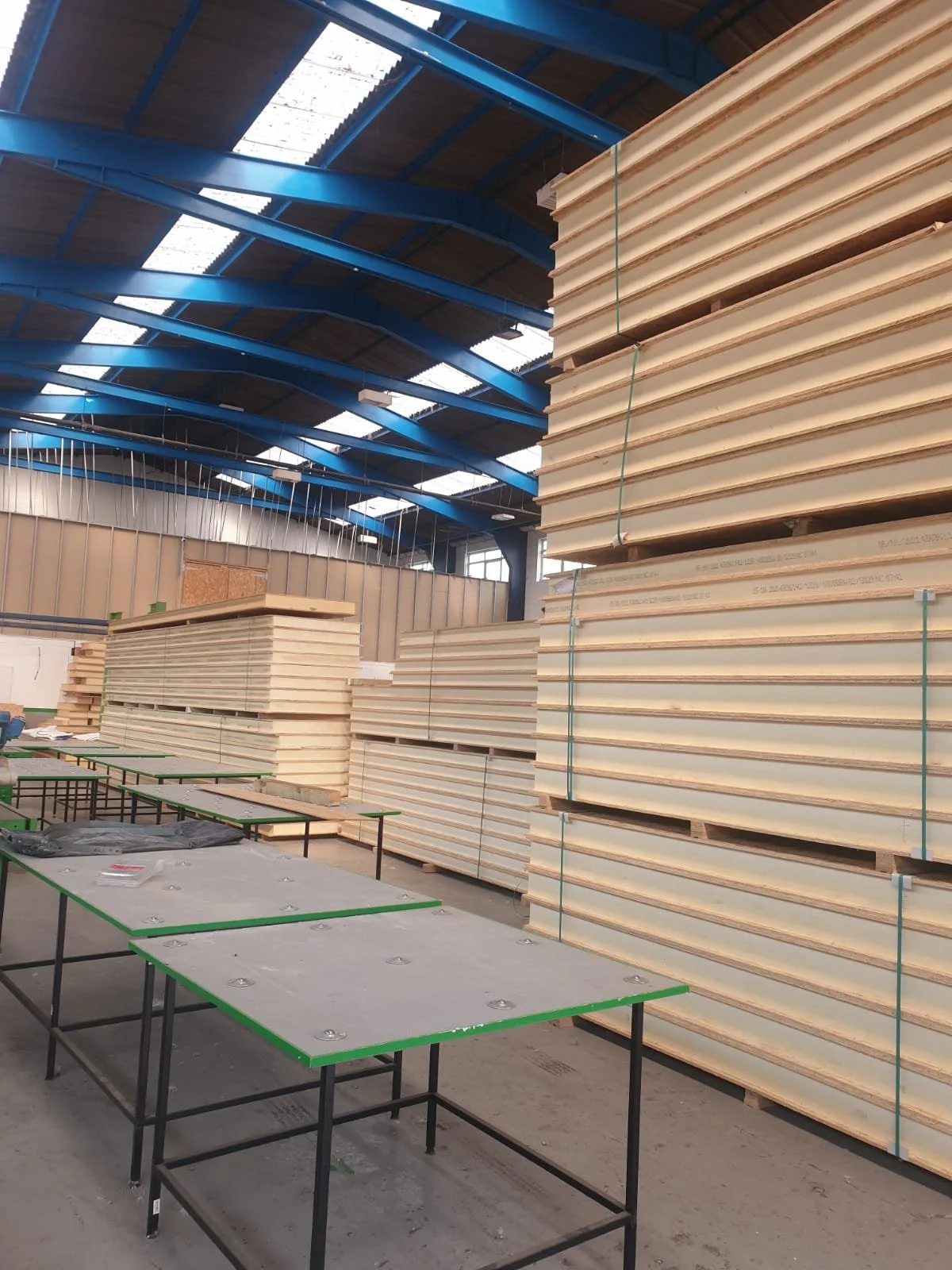 Stacked plywood sheets in an industrial warehouse with work tables in the foreground and a blue steel arched ceiling.