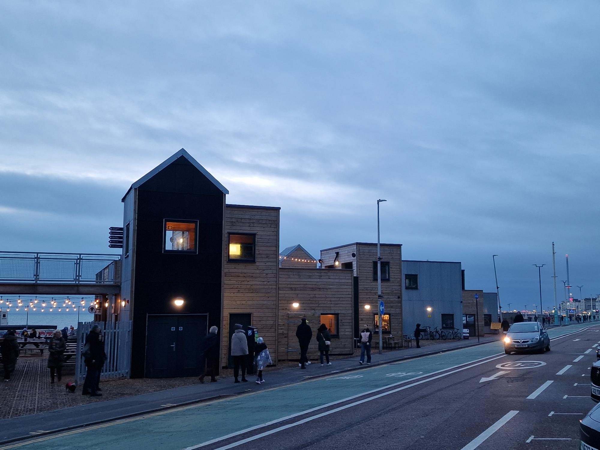 People walking along a city street near modern buildings at dusk, with a cloudy sky overhead and string lights illuminating an outdoor area.