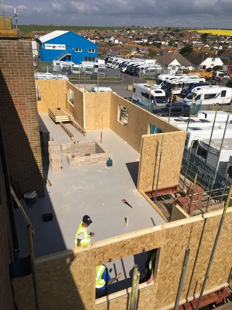 Construction site showing wooden wall panels being installed on a building's upper floor, with a worker in a yellow safety vest and helmet, in an area with parked RVs and houses in the background.