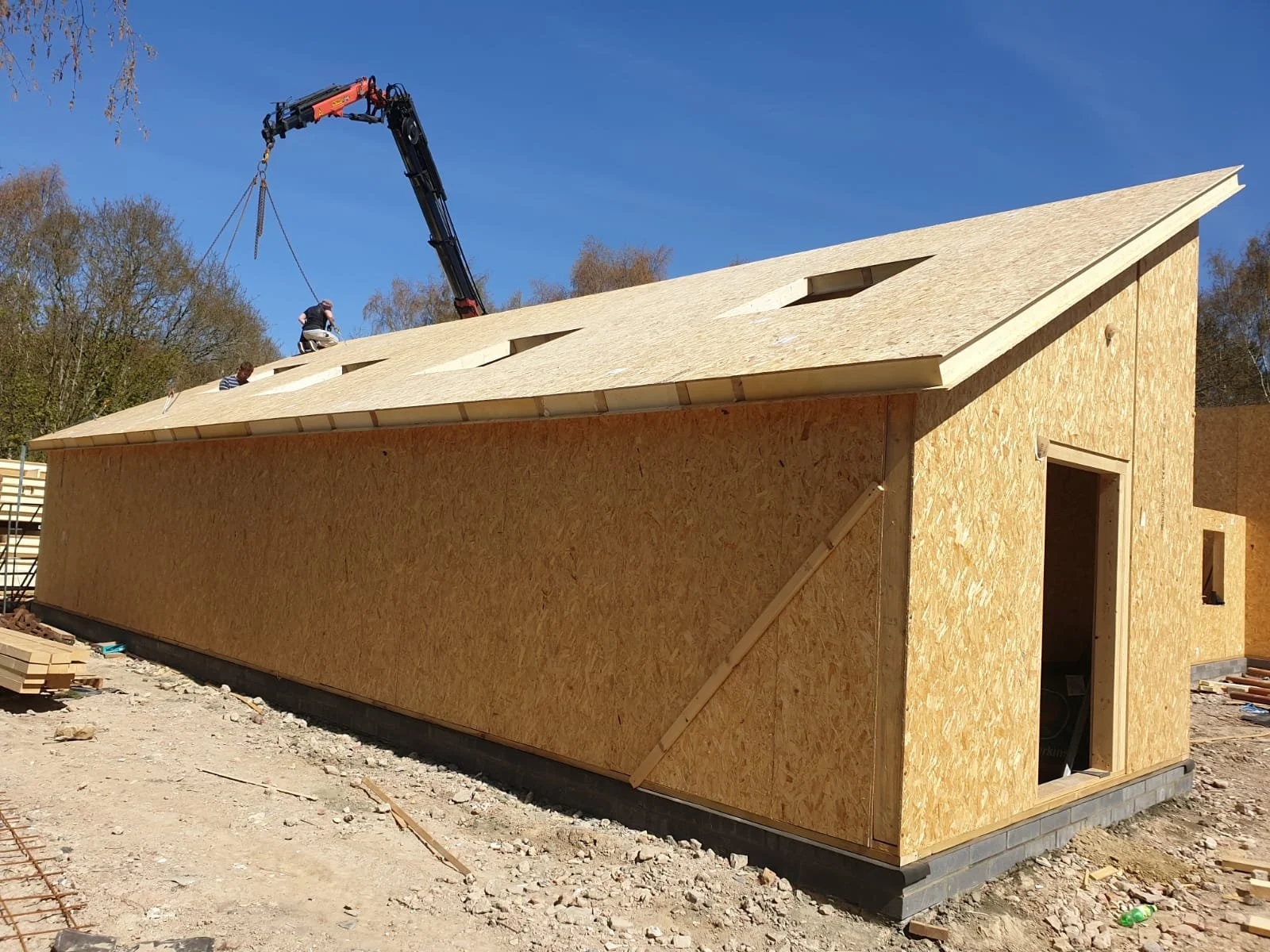 Construction site with a house in early stage, featuring wooden walls and a roof under construction with visible workers and a crane.