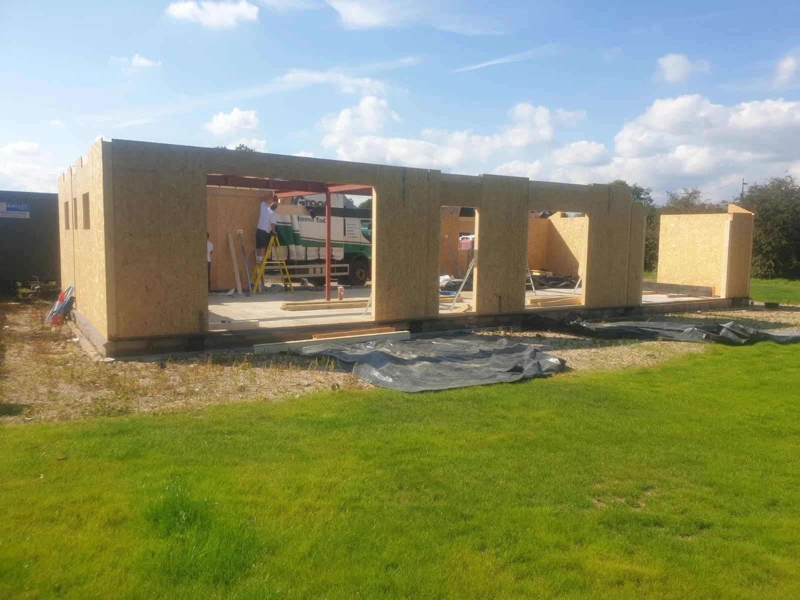 Construction of a small building with oriented strand board walls, workers assembling the structure, and a green lawn in the foreground under a partly cloudy sky.