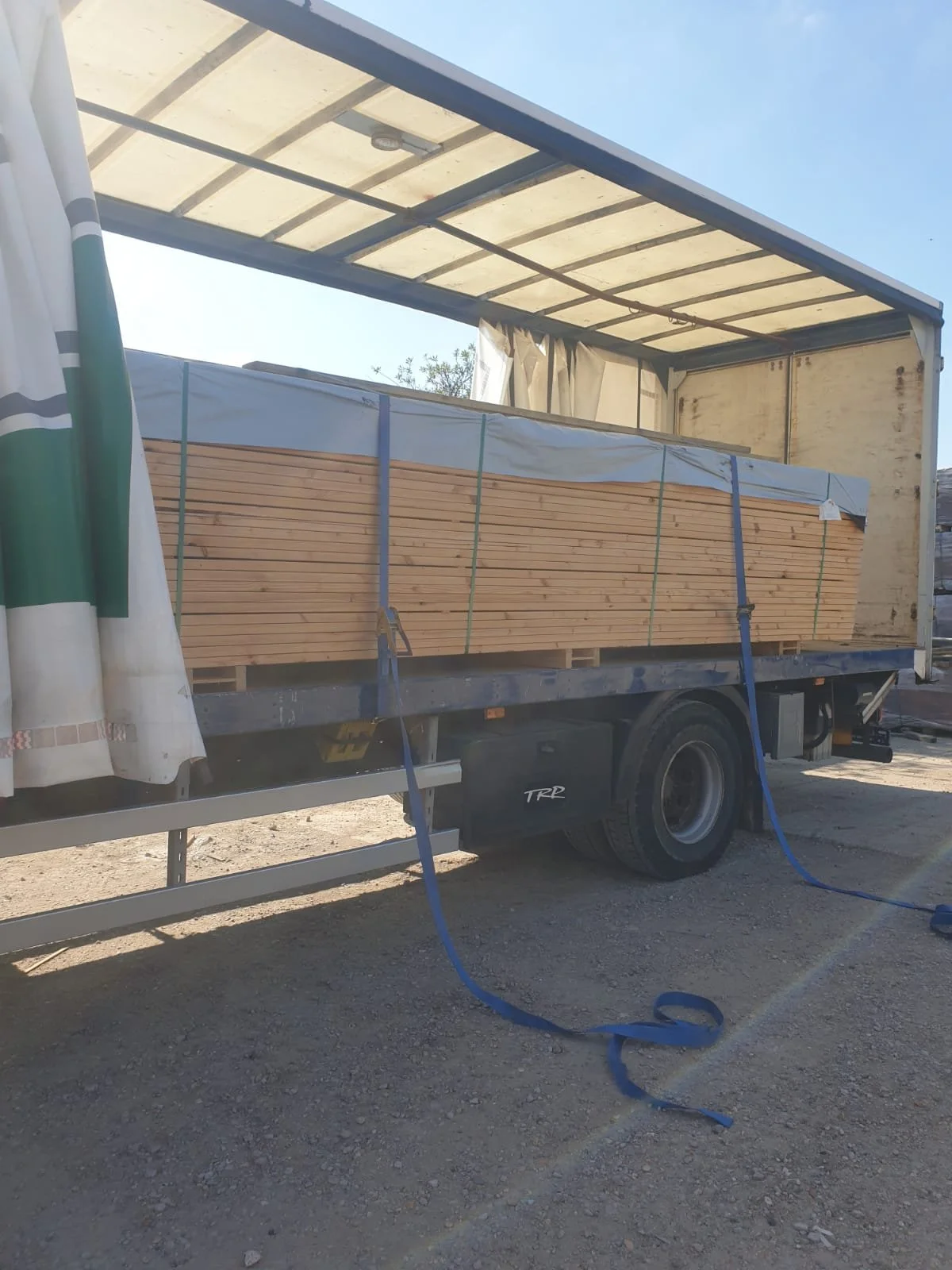 A flatbed truck with a partially open canopy carrying a large stack of wooden planks wrapped in plastic, parked on a dirt surface under a clear blue sky.