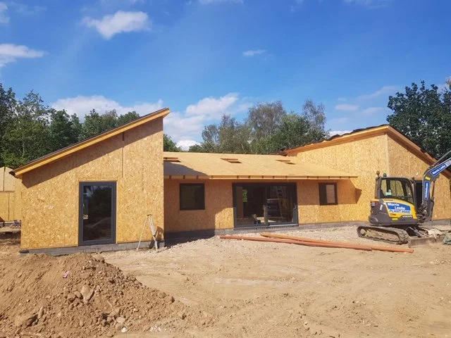 Under construction house with slanted roof panels, wooden framing, and a small excavator on site under a cloudy blue sky.