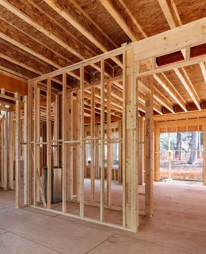 Interior view of a house under construction with exposed wooden framing and ceiling beams, and unfinished floors. There are windows showing trees outside.