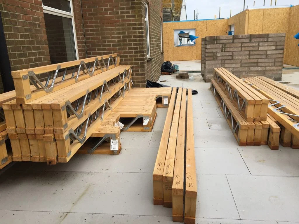 Construction site with stacked wooden benches, a partially built brick wall, and plywood barriers in the background.