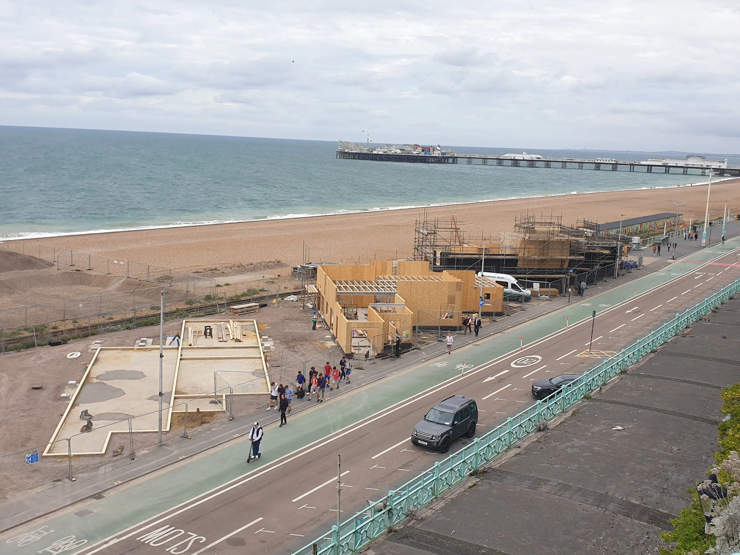 Beach with a pier extending into the ocean, some construction on the beach, and pedestrians walking along the sidewalk. Overcast sky.