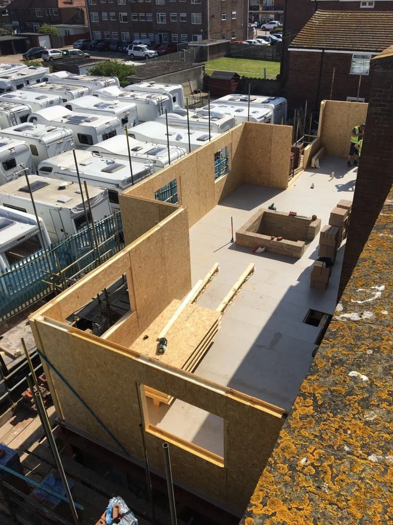 Construction site with building walls made of plywood, partial framed structure, construction tools, bricks, and workers on site, adjacent to a parking lot with many RVs.