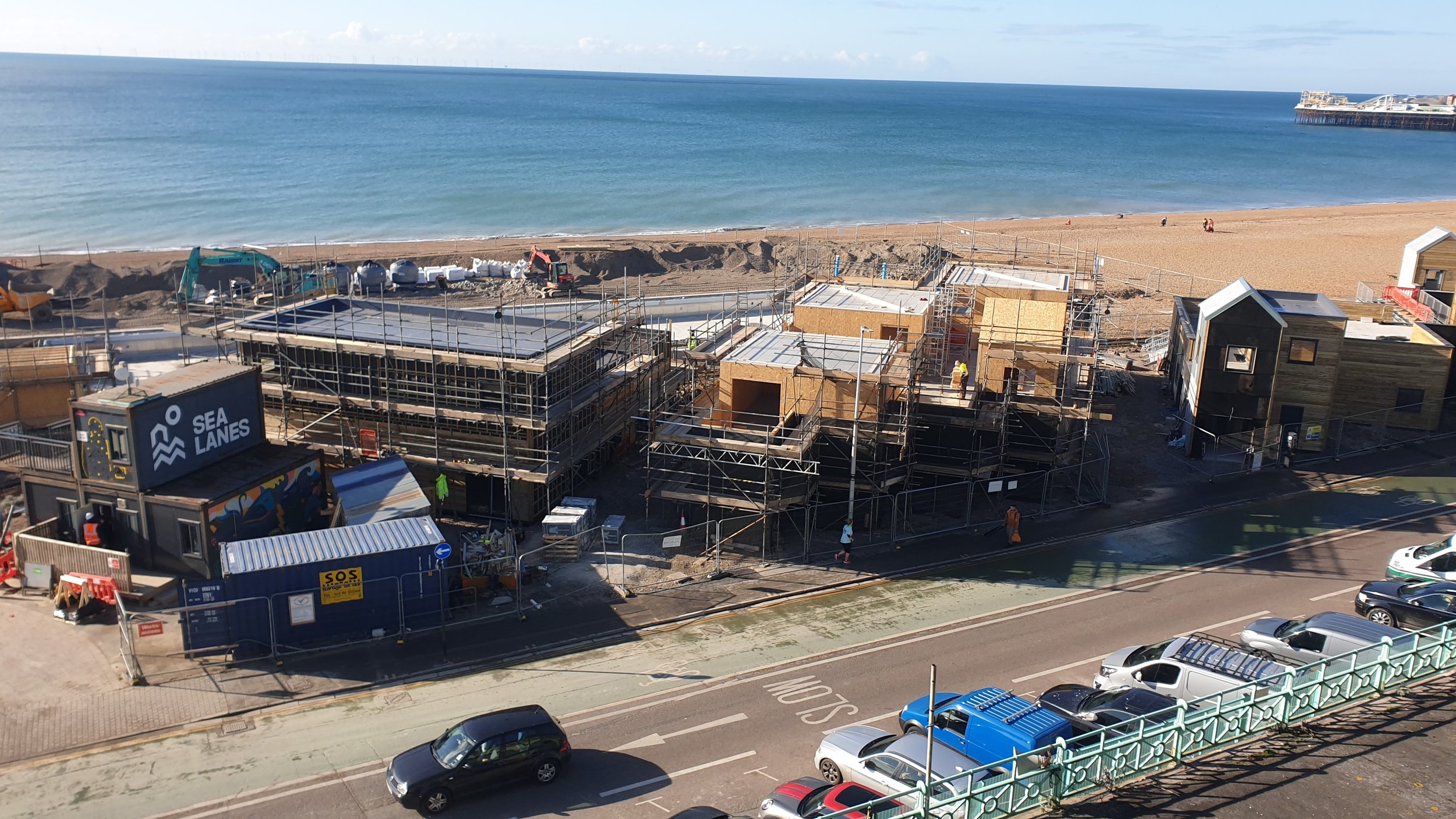 Construction site near a beach with partially built buildings, scaffolding, construction vehicles, and construction workers. The beach and ocean are visible in the background.