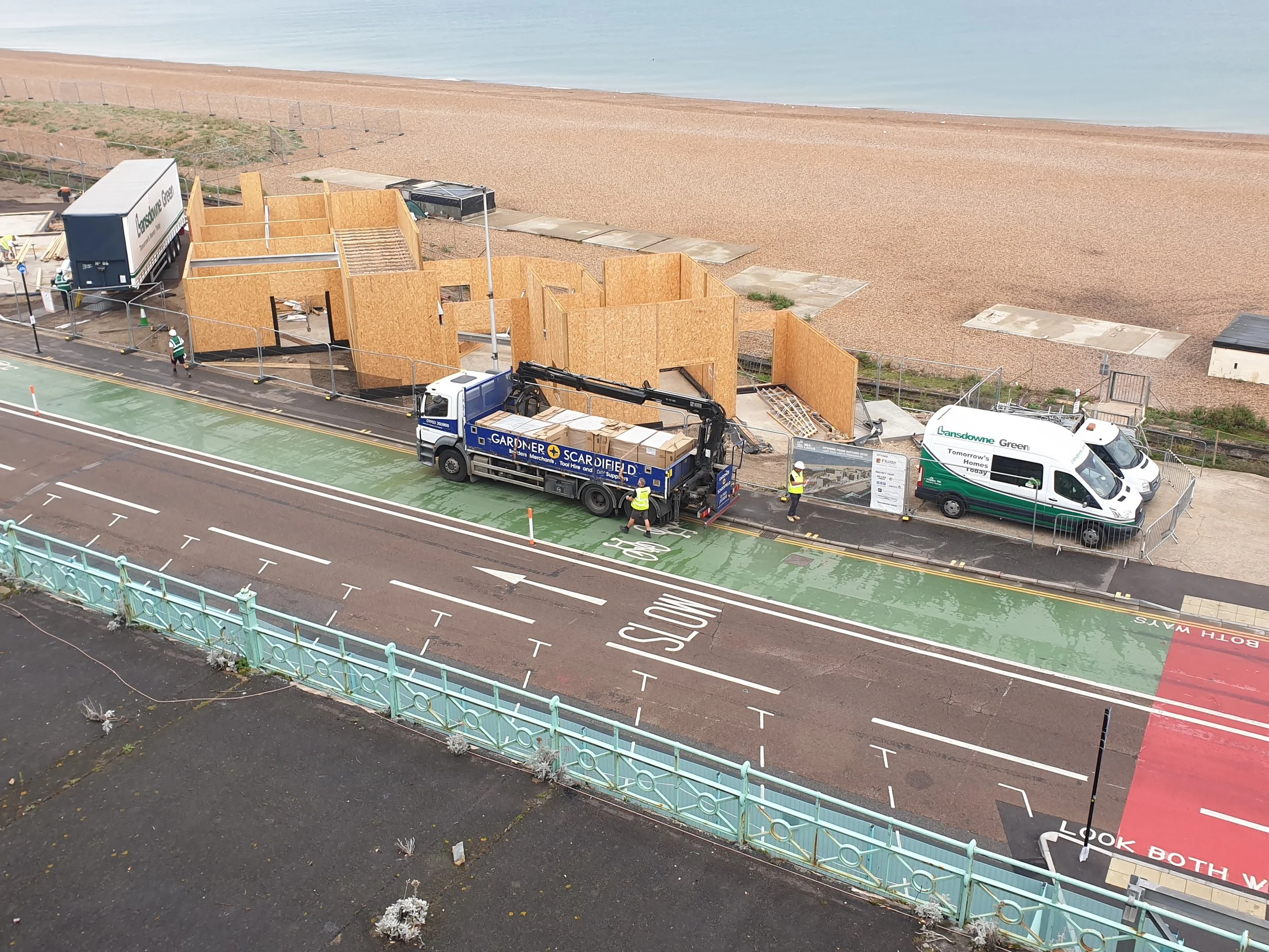 Construction site with wooden frame structures and construction workers near a beach, with trucks and a parking area in the foreground.