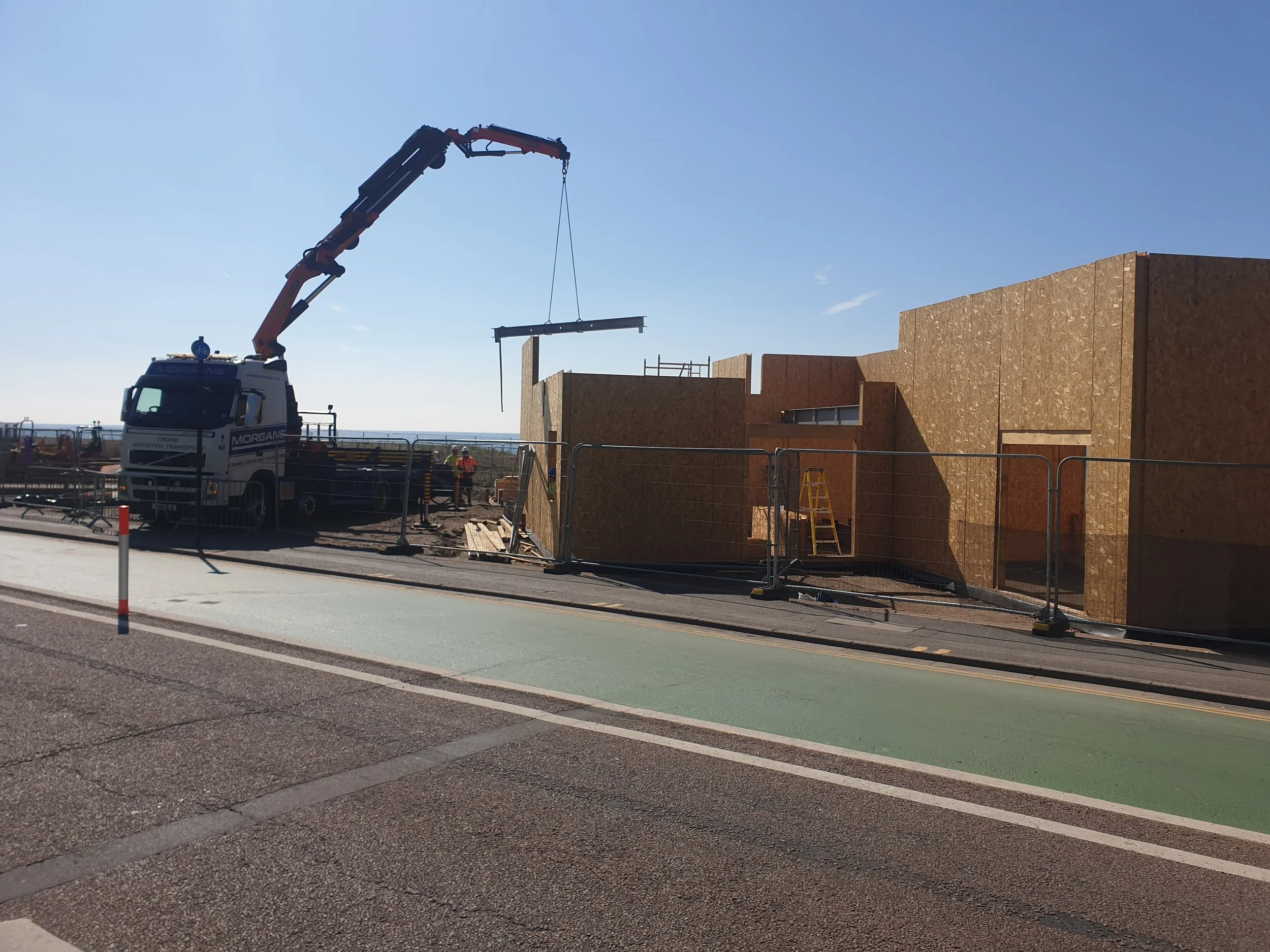 Construction site with a crane lifting a large panel of wood or similar material, construction workers present, and a building under construction with wooden exterior panels. Safety fencing and a road with green and gray markings are in the foreground.