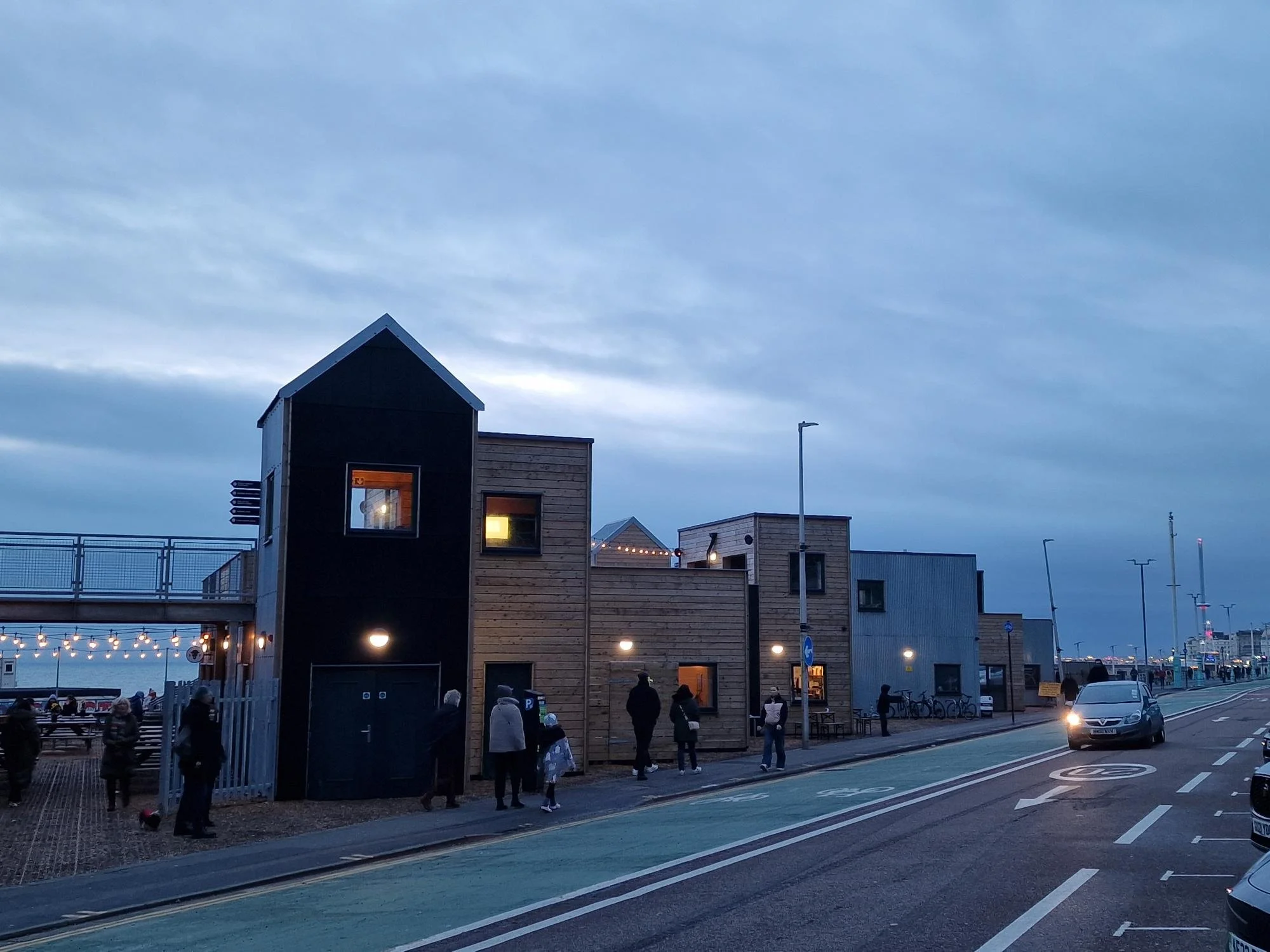 A modern urban scene with a wooden house-like structure on the sidewalk, pedestrians walking past, a bike lane, and cars on the street during dusk with a cloudy sky.