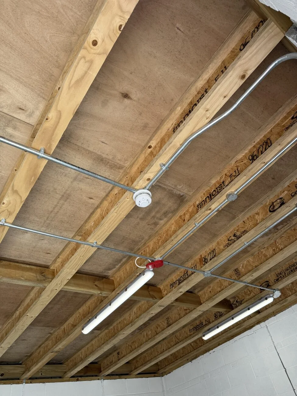 Ceiling of a building under construction with exposed wooden beams, electrical conduit, and fluorescent light fixtures.