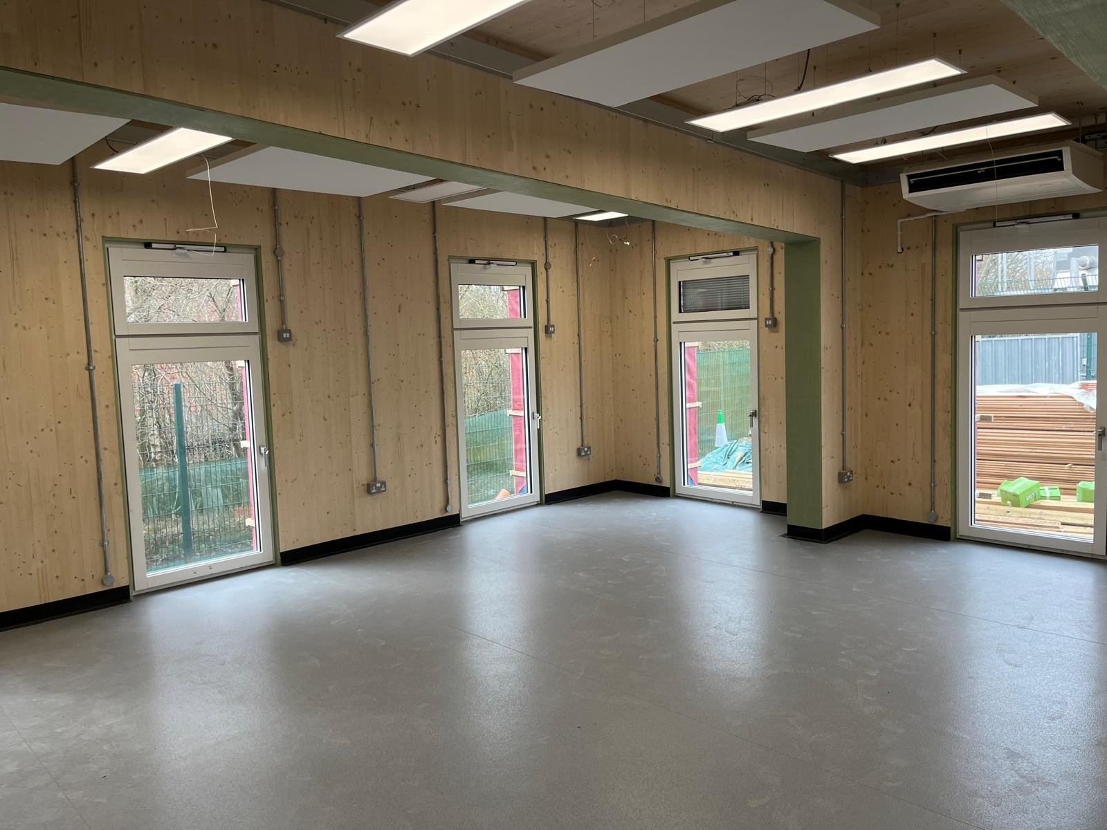 An empty room under construction with light-colored wooden walls, large windows, and ceiling panels. Outside construction materials are visible through the windows.