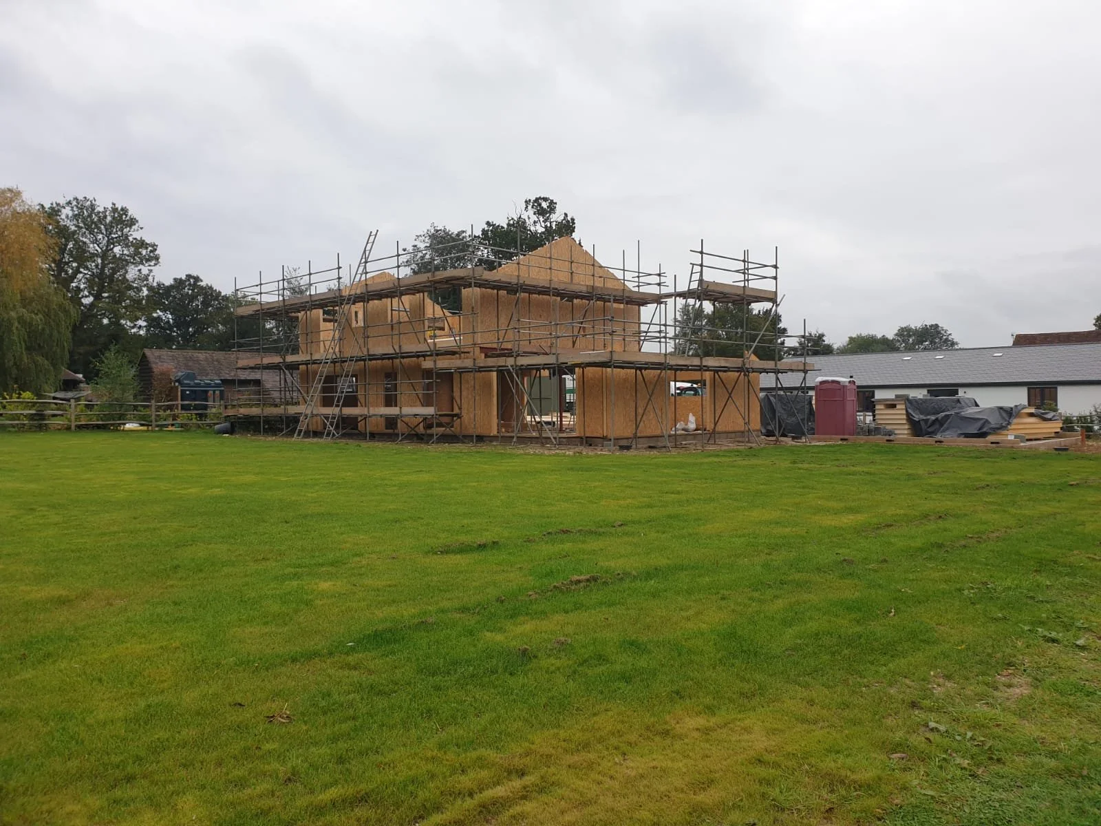 Under construction house with wooden frame and scaffolding, surrounded by green grass and overcast sky.