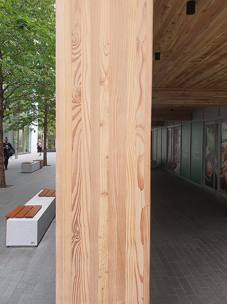 A large vertical wooden post on a city sidewalk, with trees and pedestrians in the background.