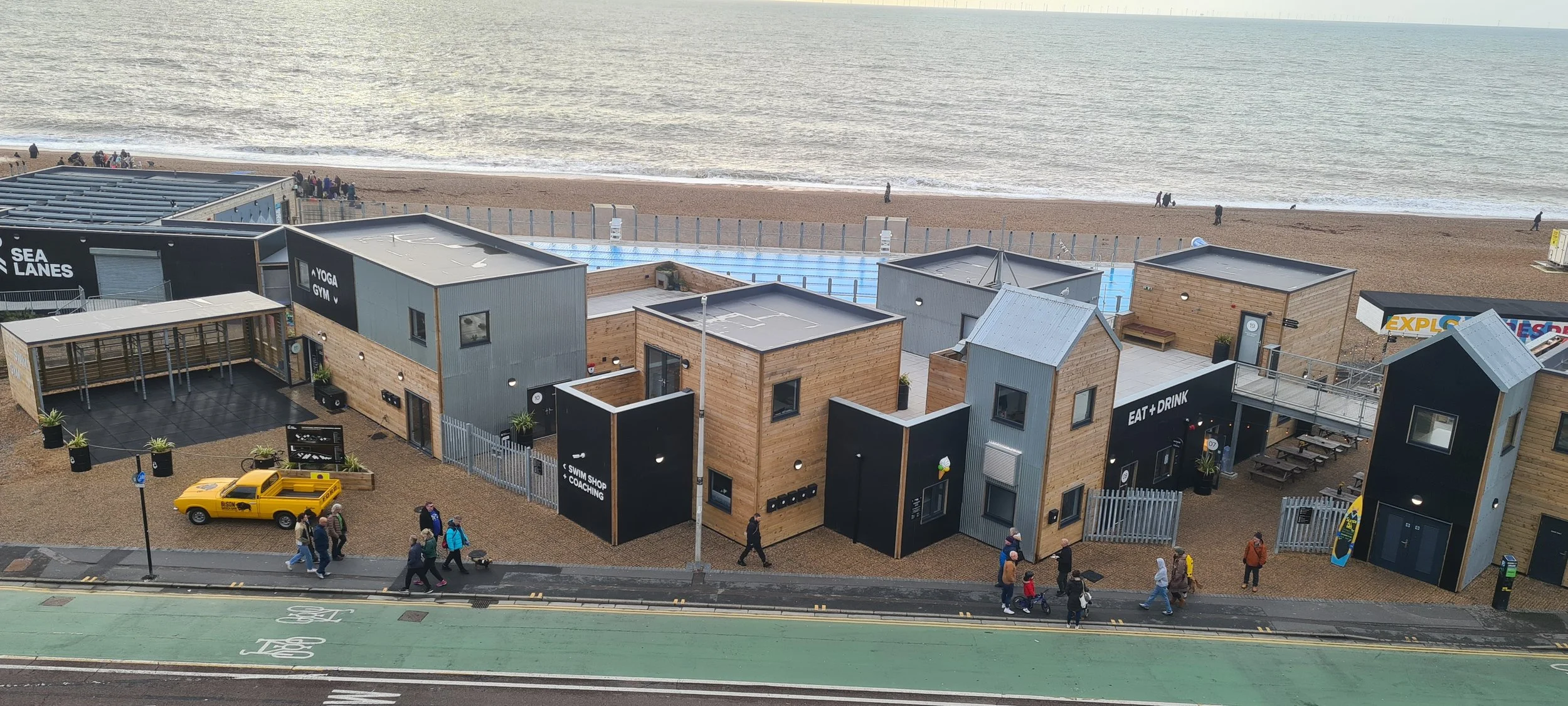 Beachfront area with modern wooden and metal buildings, including a swim shop, gym, and eat and drink establishments, overlooking the ocean with people walking along the shore and a bike lane in the foreground.