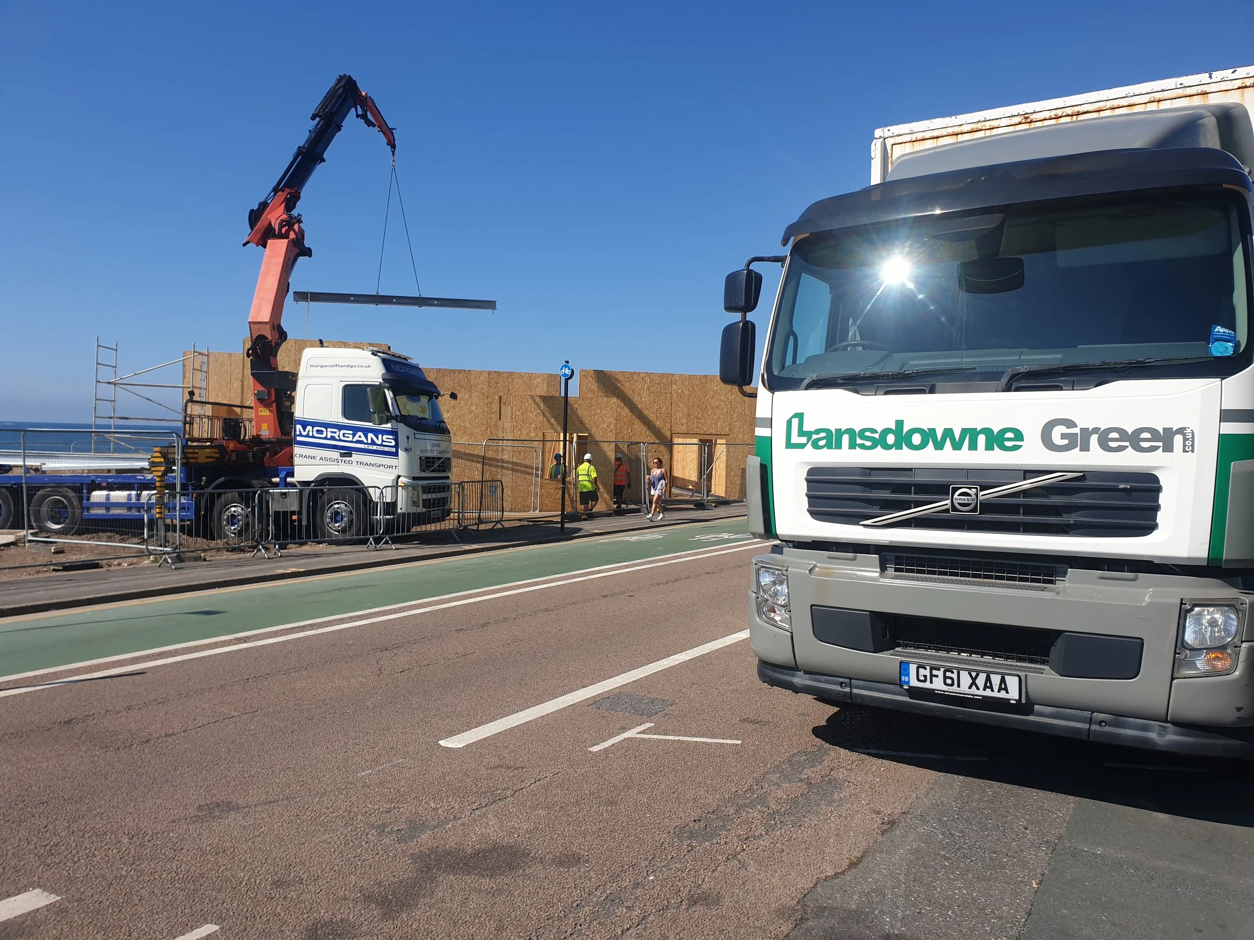 Construction site with workers, a crane lifting a large wooden panel, and trucks nearby under a clear blue sky.