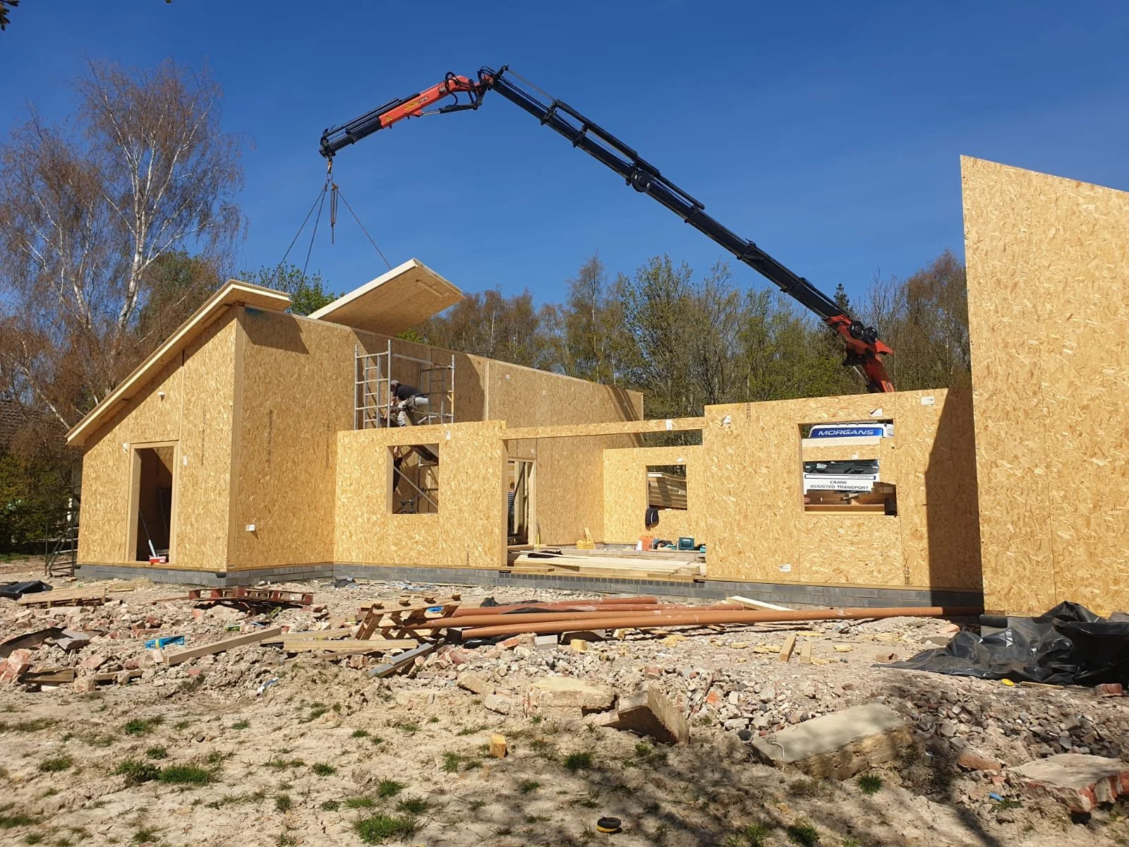 Construction site of a house with wooden walls and a crane lifting a large wooden panel, workers installing parts, and scattered building materials on the ground under a clear blue sky.