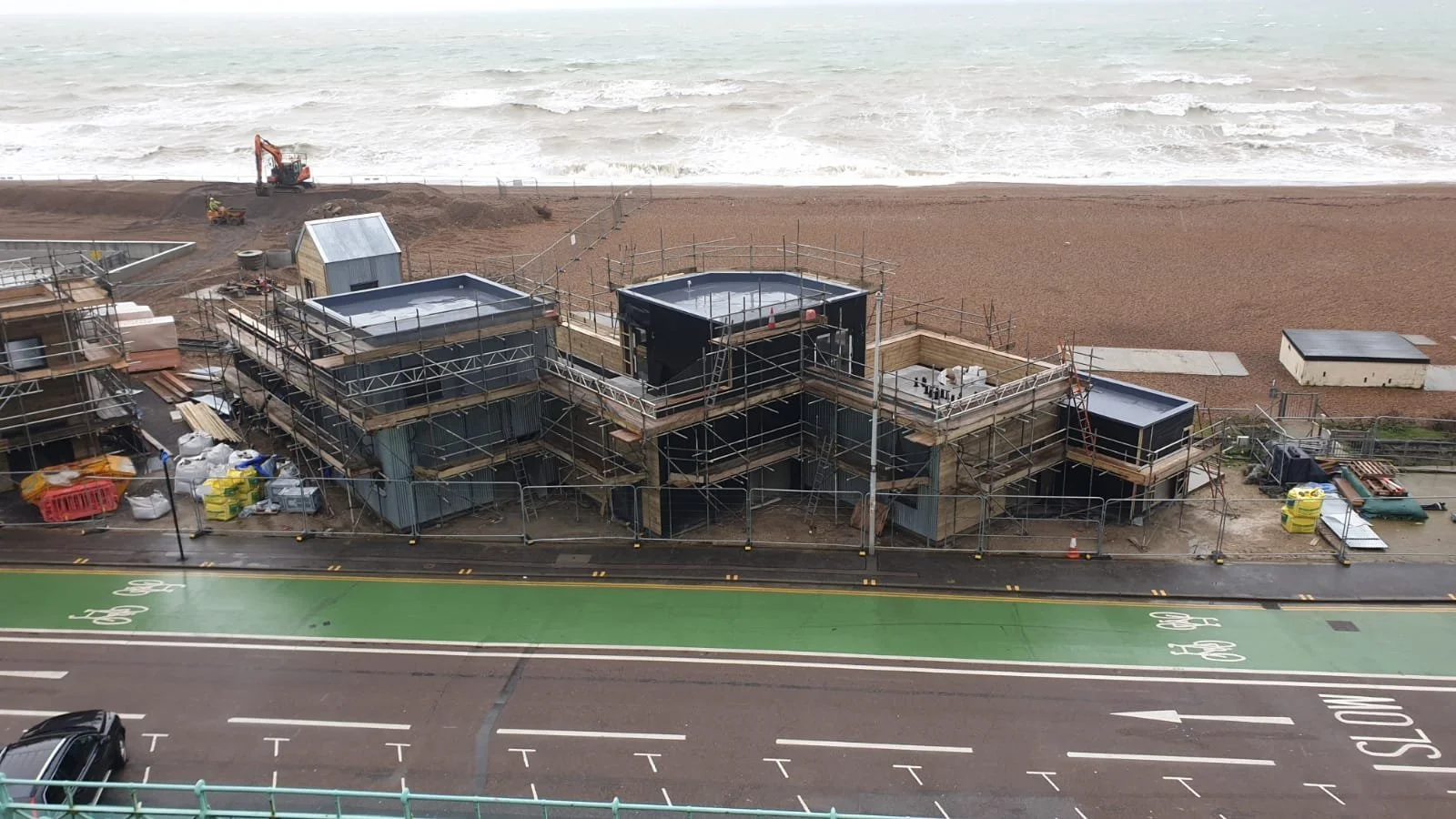 Construction site by the beach with scaffolding around buildings, construction materials, and equipment. The beach and ocean are visible in the background.
