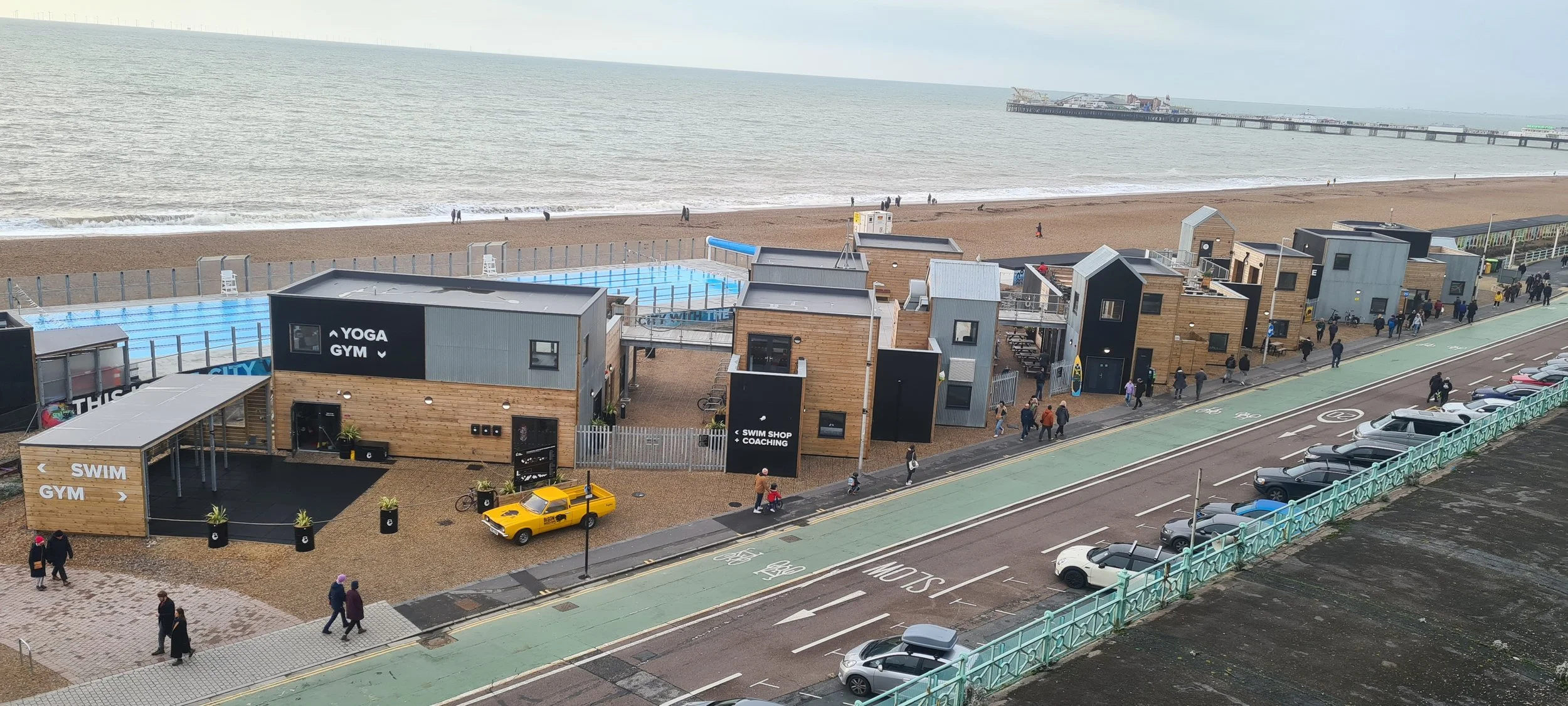 Beachside area with wooden buildings, a swimming pool, and people walking along the sidewalk. There are parked cars and a bike lane on the road, with the ocean and a pier in the background.