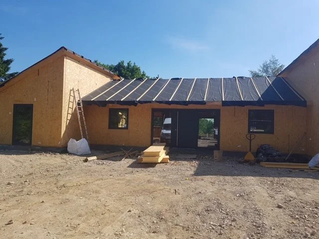 Under construction house with plywood walls, a sloped metal roof, a ladder leaning against the side, and building materials on the ground.