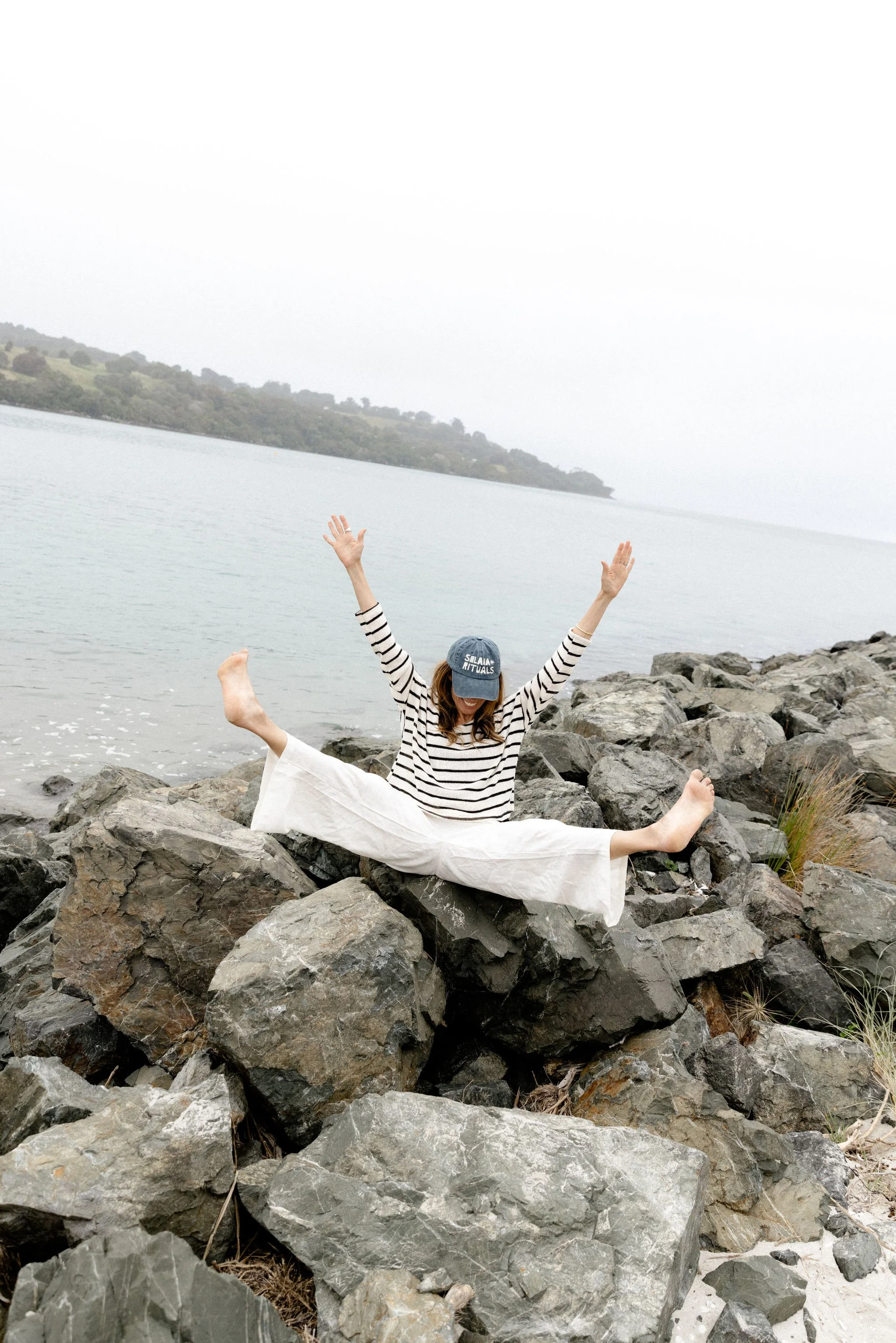 Person in striped shirt and white pants sitting on rocks by the water, raising arms in a joyful pose with arms and legs extended, wearing a cap, with a scenic background of water and land.