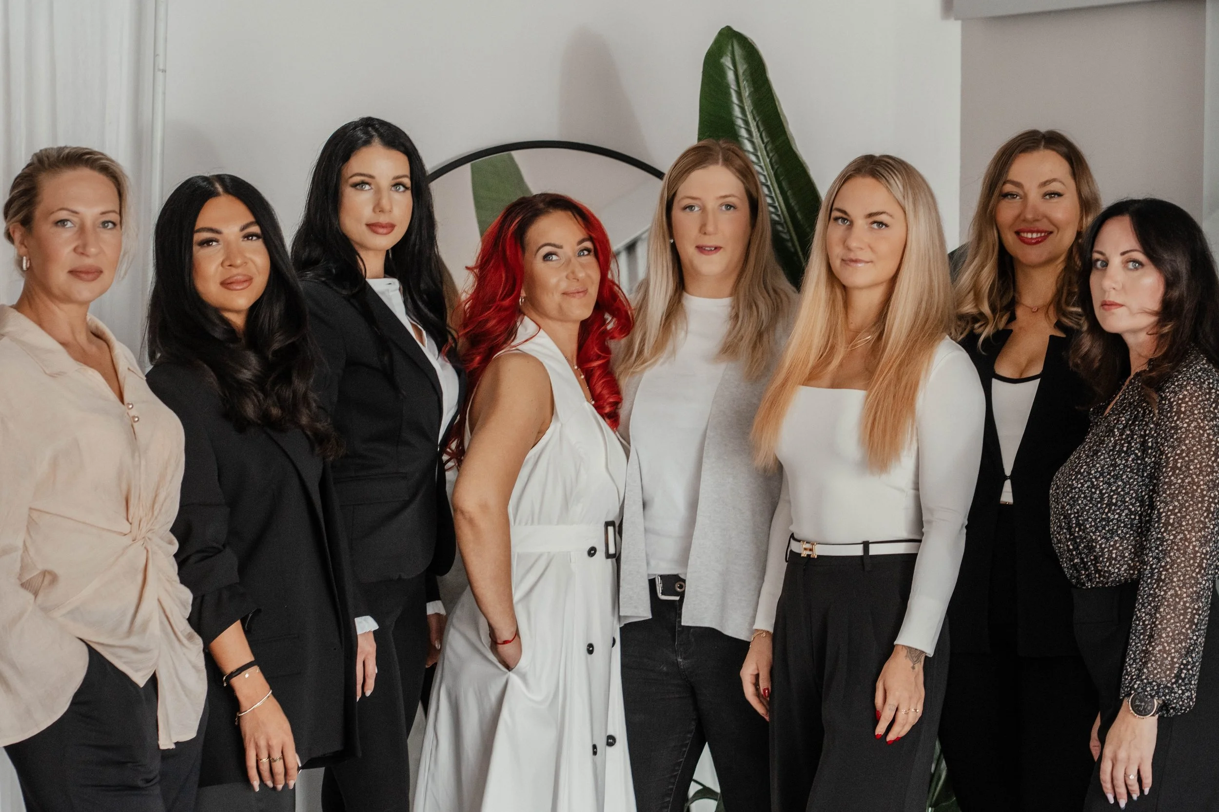 Gruppe von acht Frauen steht in einem Raum vor einem großen grünen Blatt an der Wand, lächelnd, für ein Foto posierend.