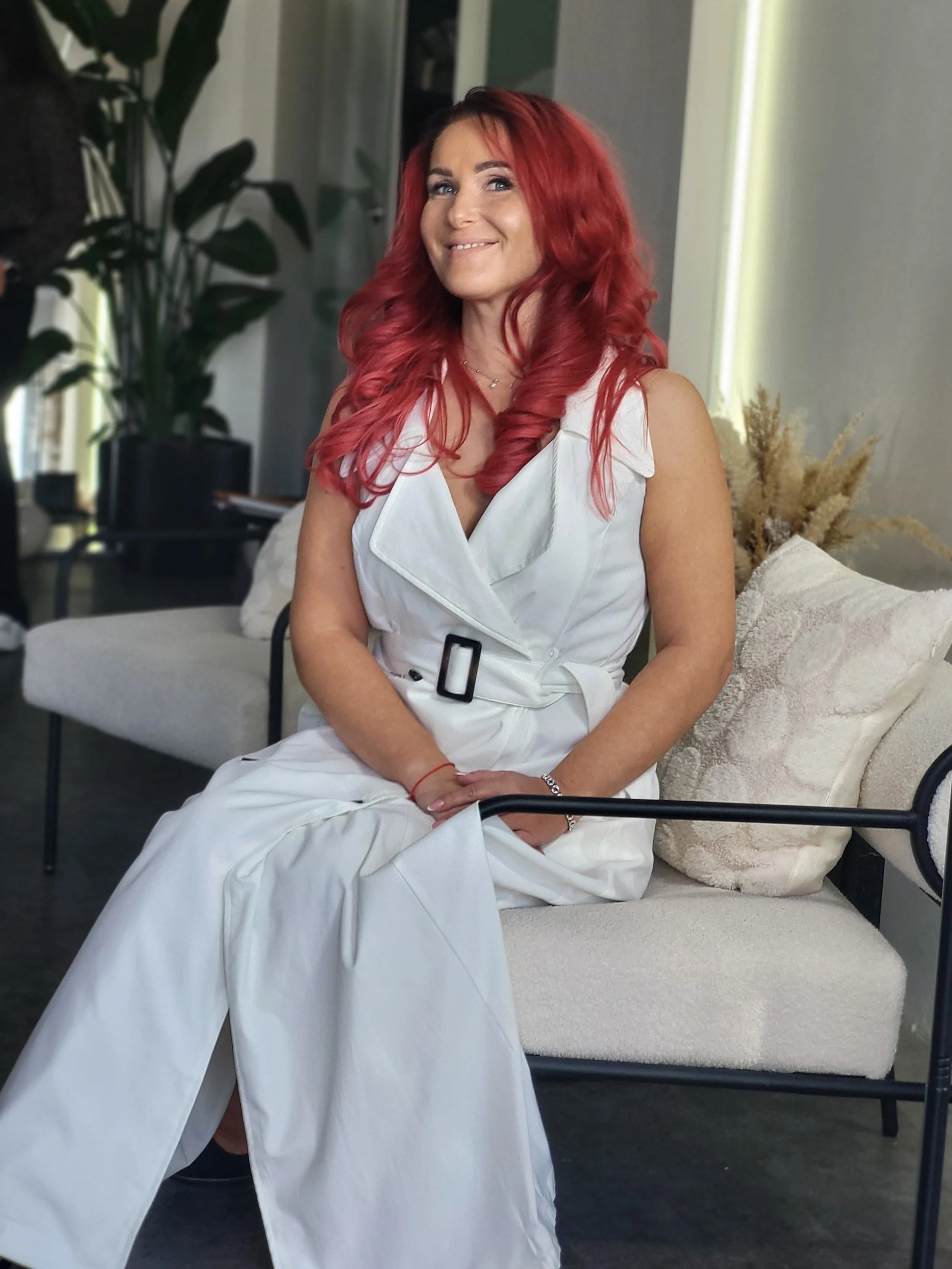 Eine Frau mit roten, lockigen Haaren sitzt auf einer weißen Couch in einem modernen Raum, trägt ein weißes Kleid und lächelt.
