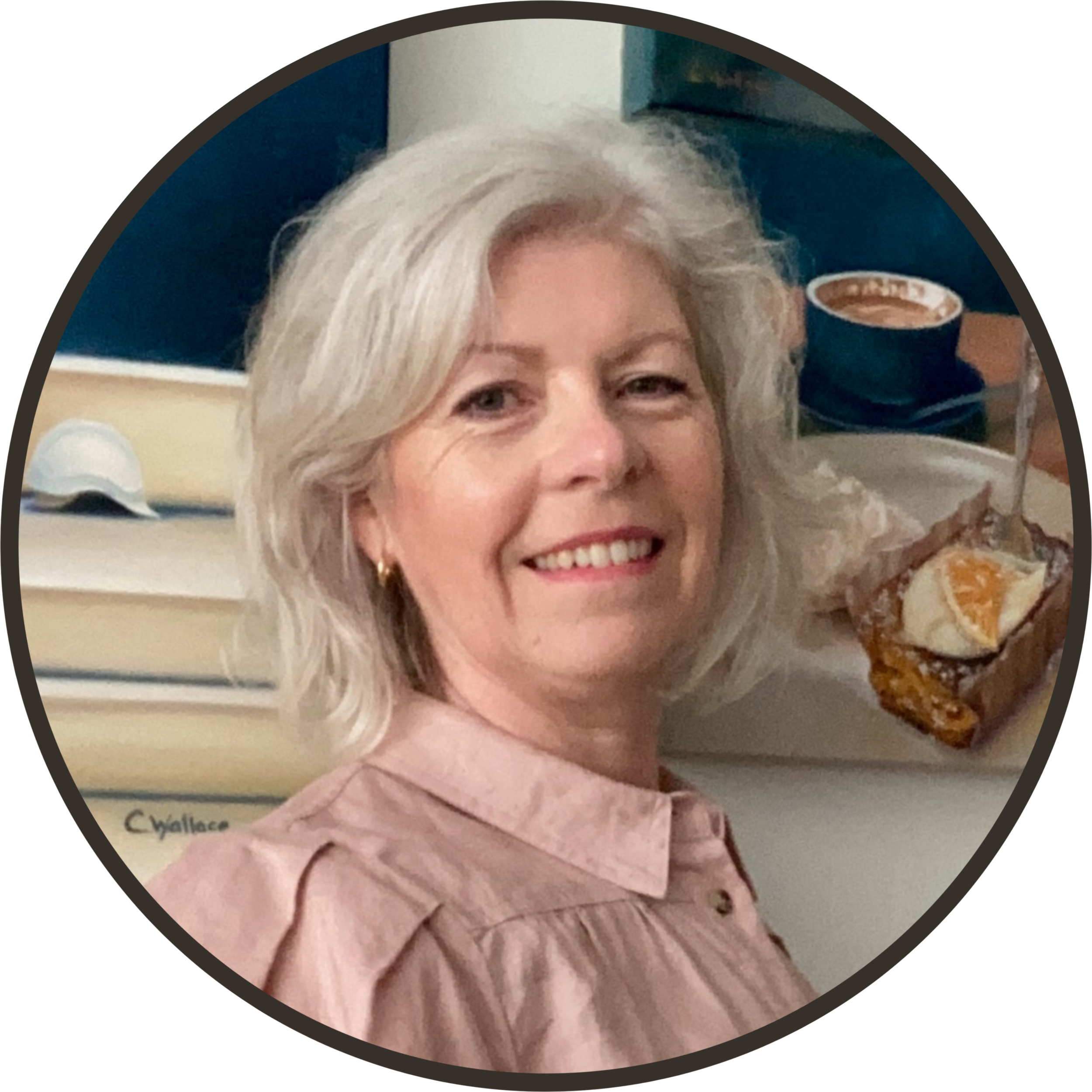 A woman with blonde hair smiling, sitting in an office or workspace with a computer, stationery, and a pastry with whipped cream and orange slice on a plate.