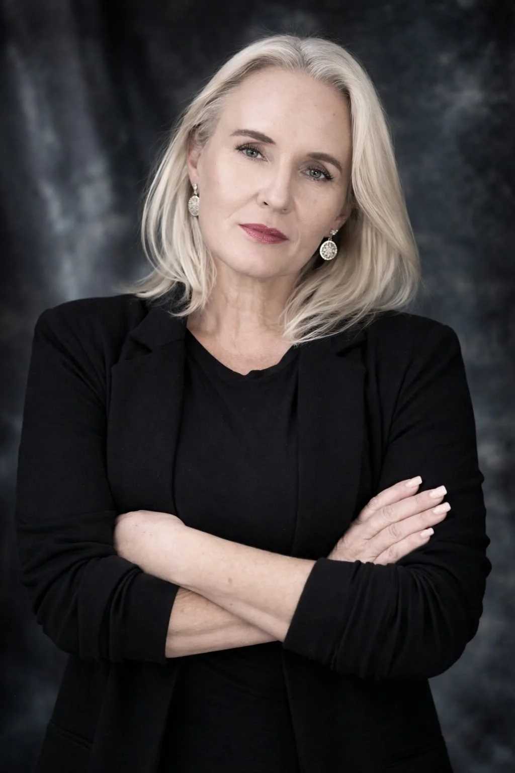 A woman with blonde hair, wearing a black blazer and earrings, posing with crossed arms against a dark cloudy background. Photography by Riaan Swiegelaar