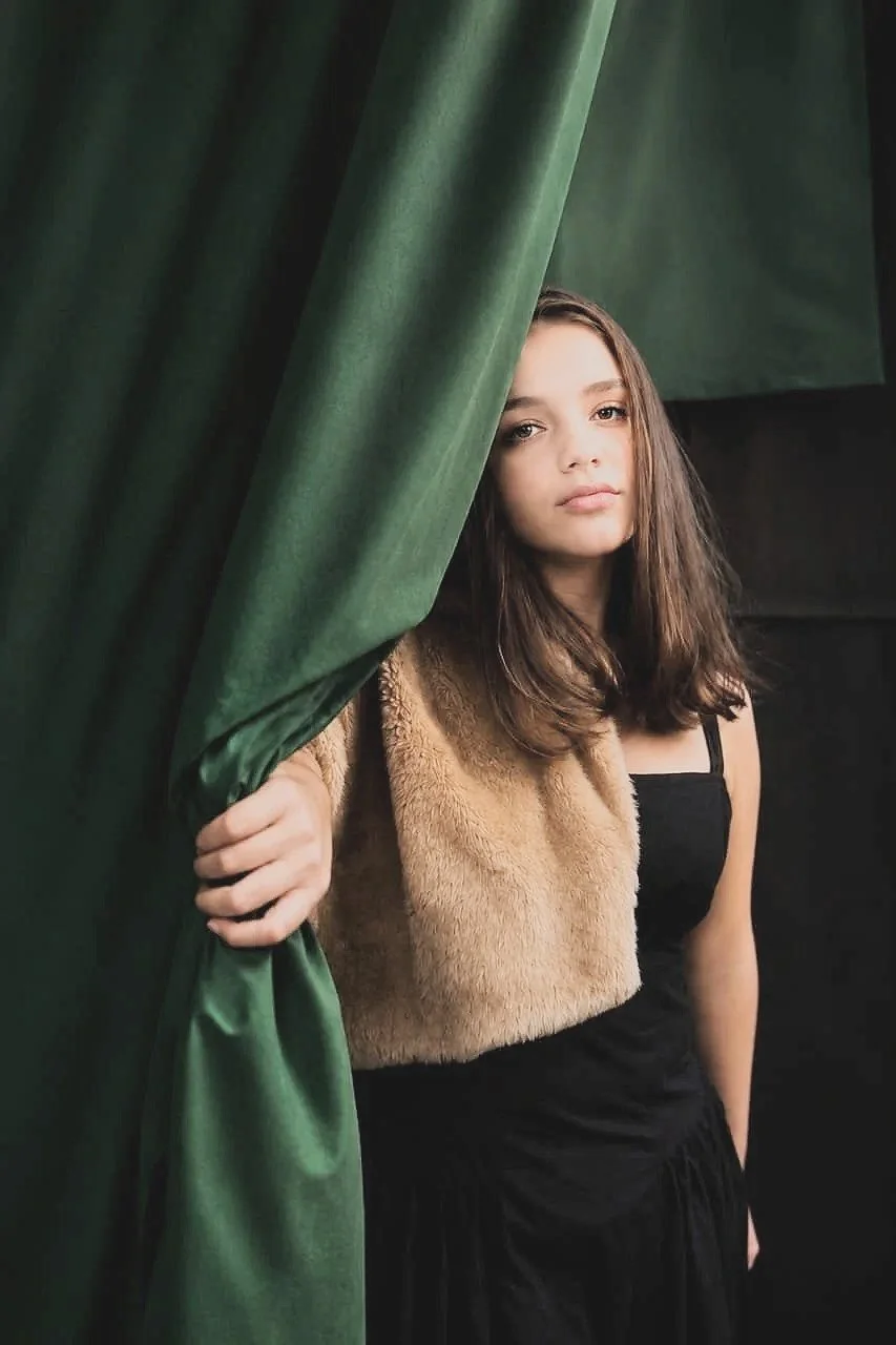 Young woman with brown hair standing partly behind green curtains, wearing a black dress and a tan faux fur vest, looking at the camera. Photography by Riaan Swiegelaar
